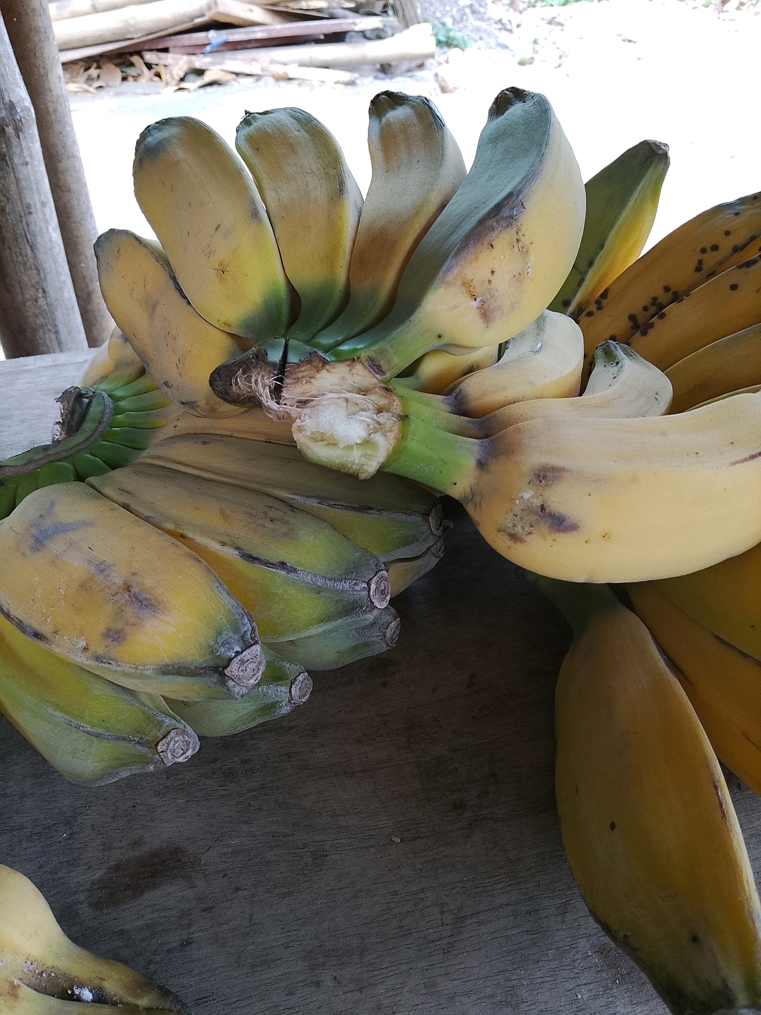 A close-up, high-angle shot shows a cluster of ripe bananas, some with yellow and green skins, on a weathered wooden surface. Several of the bananas exhibit brown spots and blemishes. The background is blurred, with hints of natural elements like wood and foliage, suggesting an outdoor setting in Banaran, Indonesia. The scene is static, with no people or apparent actions.