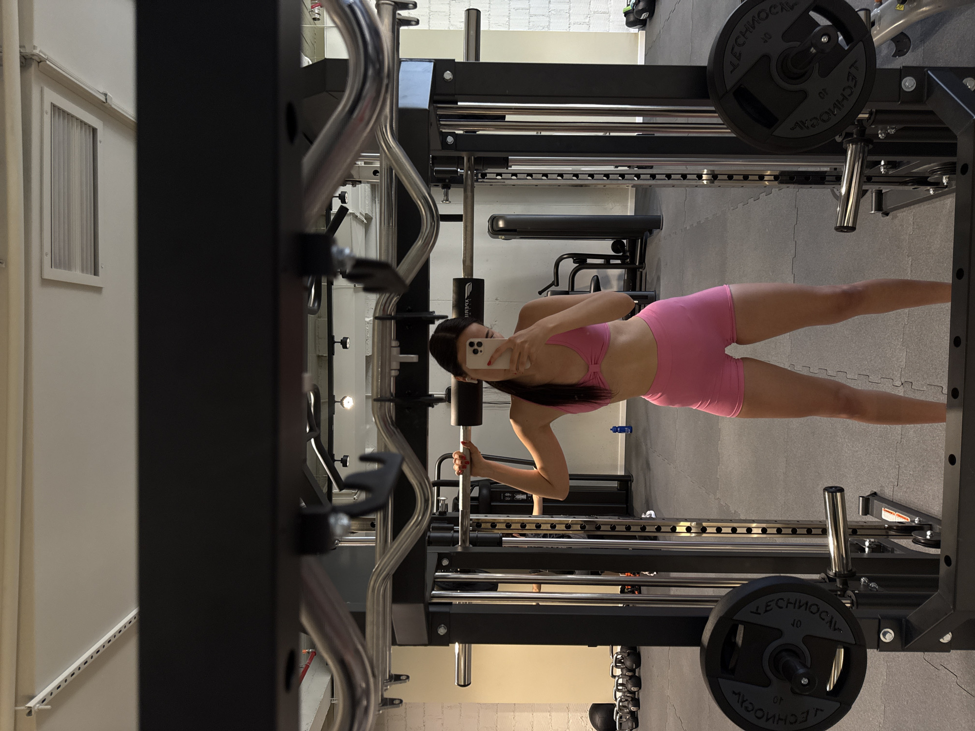 A woman in pink athletic wear is holding a smartphone in her right hand to take a picture of herself in a gym setting. The gym has a Smith machine and various weight plates. The floor is covered in gray interlocking rubber tiles. A bench and dumbbell rack are visible in the background. A window and some ventilation ducts are on the left side of the image.