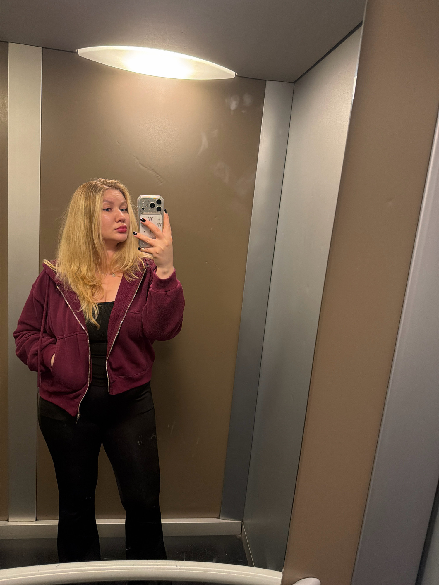 A young woman with blonde hair is taking a mirror selfie in what appears to be an elevator. She is wearing a maroon zip-up hoodie over a black tank top and black flared pants. Her left hand is in the pocket of her hoodie. The elevator walls are a combination of metallic silver and a warm brown tone. A white light fixture is visible on the ceiling. The image is taken from a slightly low angle, looking up at the woman. The overall environment is clean and modern, suggesting an urban setting. While the image does not provide explicit visual cues to confirm the exact location, the context of a modern elevator suggests it could be in a public building or transit area within the city of Koszalin, Poland.