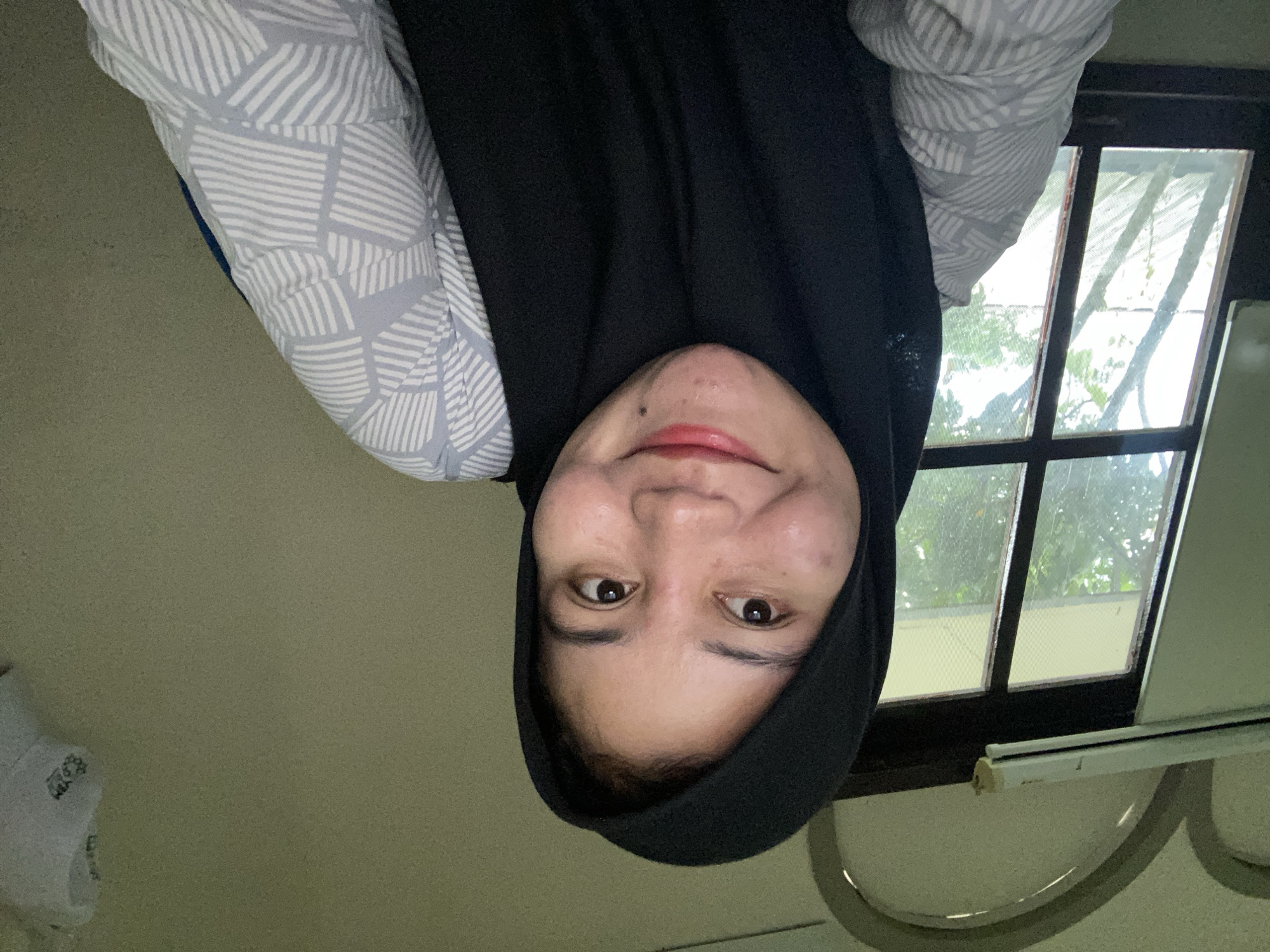 A young woman wearing a black hijab and a grey and white patterned shirt smiles at the camera. She is sitting indoors in front of a window that looks out onto trees. The room appears to be a bedroom or living area, with a light-colored wall and a ceiling with arched features. A fluorescent light fixture is visible above the window. There is also a white object with writing on it hanging on the wall to the right of the frame. The overall impression is of a casual, indoor setting in a residential area of Banjarbaru, Indonesia.