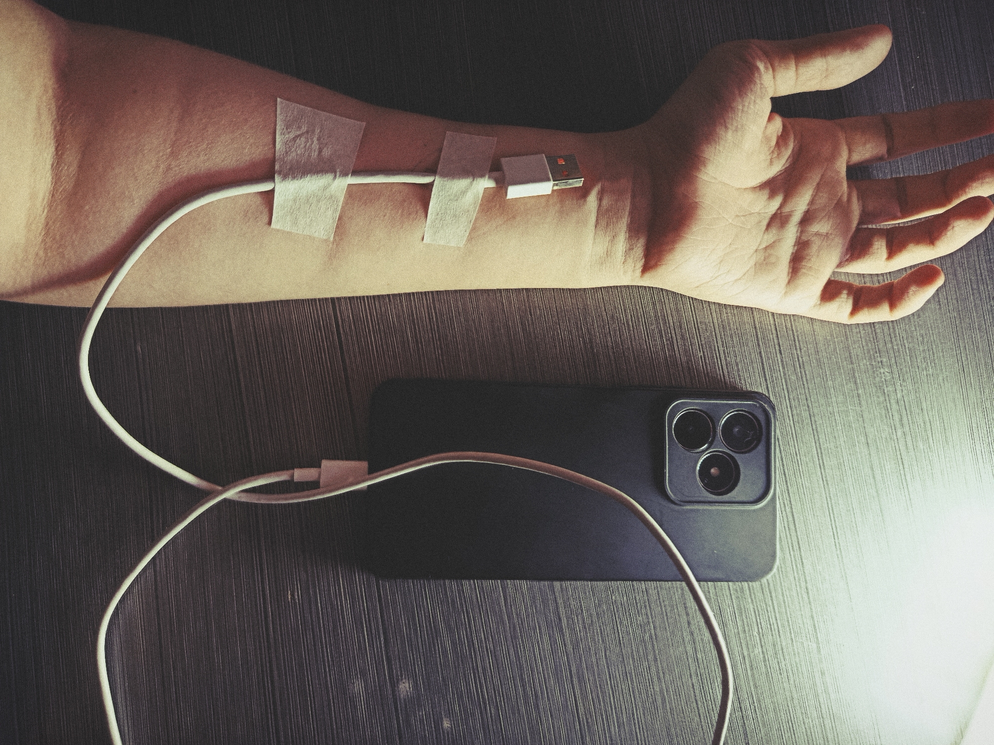 The image presents a human forearm and hand resting on a dark, textured surface, likely wood. A white USB charging cable is taped along the forearm using three strips of light-colored adhesive tape, resembling a medical intravenous line. The USB-A end of the cable is positioned near the wrist, appearing to "inject" into the arm.
The other end of the charging cable is connected to a black smartphone, visible lying face down on the same surface next to the arm. The phone features a distinct triple-camera array on its back.
The scene is dimly lit, with a strong light source originating from the right side, casting prominent shadows and highlights across the hand, arm, and part of the smartphone. The overall aesthetic is slightly desaturated and grainy, contributing to a somber or reflective mood.
The activity depicted is not literal but symbolic: a human arm is "charged" by a smartphone, illustrating a profound dependence on technology, akin to a life-sustaining medical procedure. This visually communicates themes of addiction to or vital integration of smartphones into human life.
There is no visible text in the image. The image effectively uses visual metaphor to convey its message.