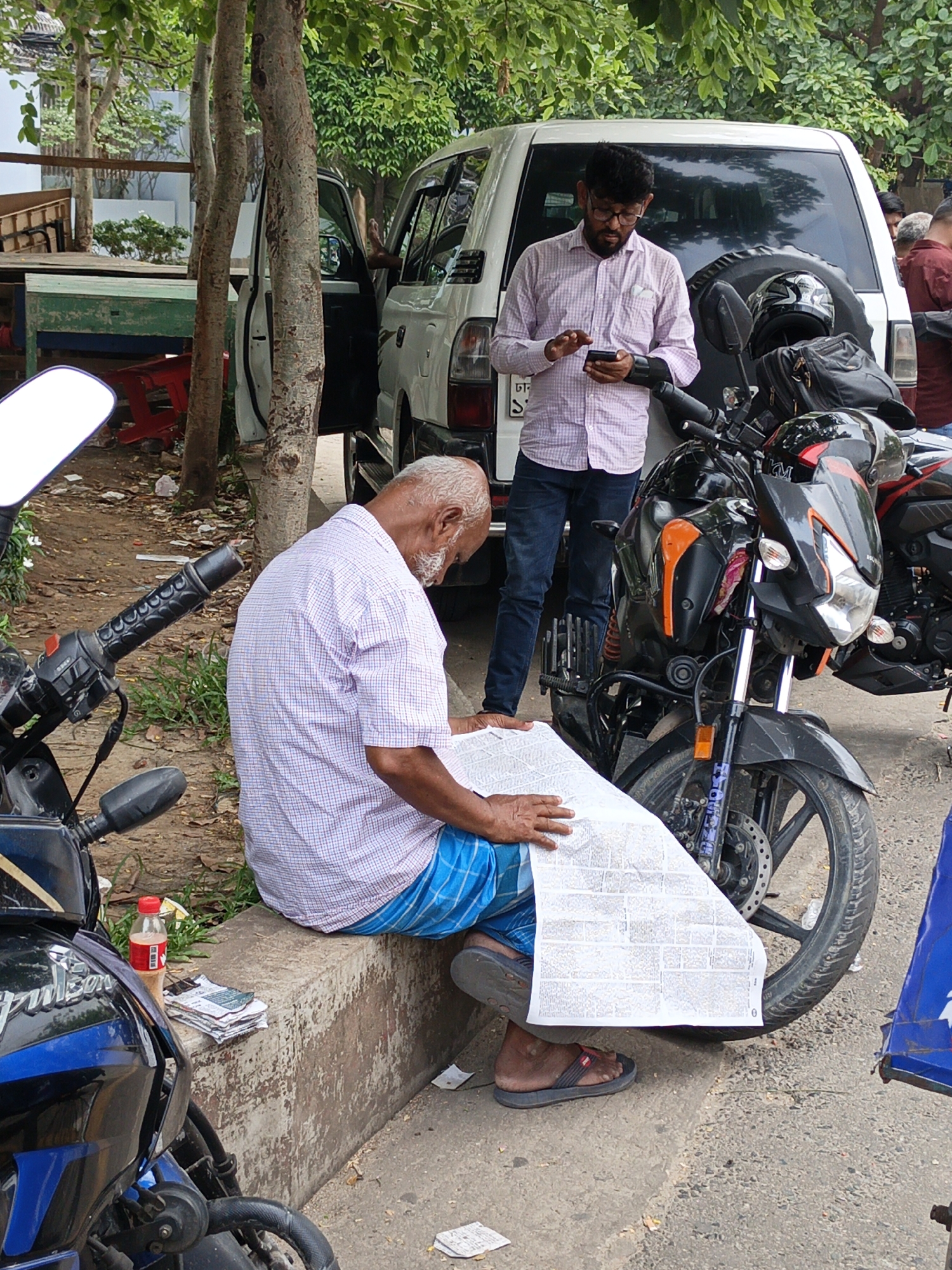 The image shows an elderly man sitting on a low concrete barrier reading a large newspaper. He is wearing a short-sleeved, light-colored, checkered shirt and blue checked shorts. He has gray hair and wears sandals. To his left is a motorcycle, next to a coca-cola bottle. To his right, a black motorcycle is parked. A stack of what appears to be newspapers sits on the barrier near his feet.
Behind him, a man wearing glasses, a light pink patterned shirt, and jeans stands looking at his phone, next to a white vehicle with the door open. The environment seems to be an outdoor urban setting with trees in the background.