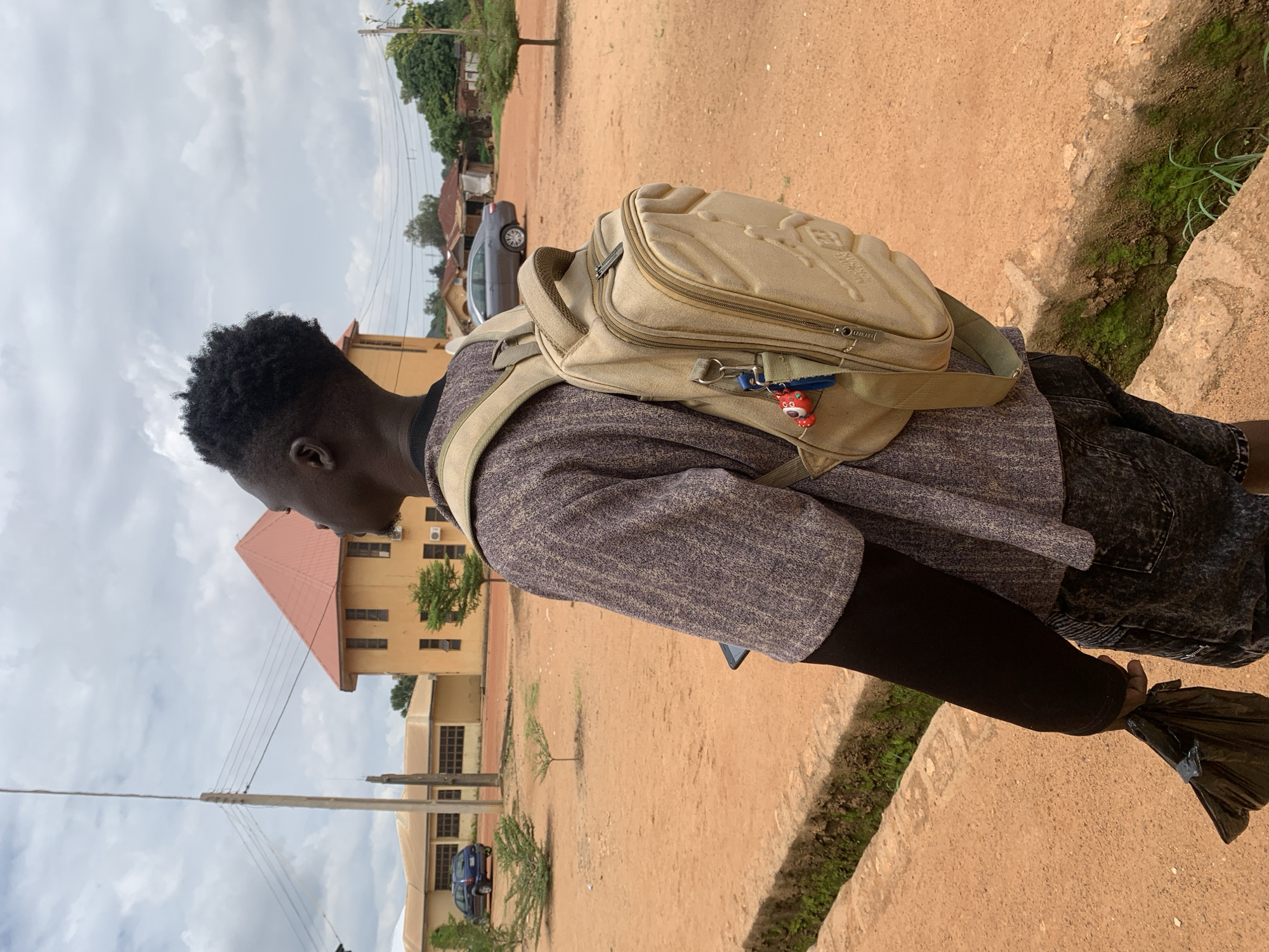 The image captures a male individual from behind, standing on reddish-brown, dusty ground under an overcast sky, in what appears to be a non-paved area of Jos, Nigeria. He wears a short-sleeved, patterned greyish-purple shirt over a long-sleeved black undershirt and dark, patterned shorts. A tan-colored backpack, featuring an embossed "Jordan" logo on its main compartment, is worn on his back, with a set of keys and a red character keychain attached to its left strap. A dark object, possibly a smartphone, is visible in his left pocket, and he holds a crumpled black plastic bag in his left hand. A concrete drainage ditch with green moss and sparse vegetation runs along the immediate foreground. In the mid-ground, a tall utility pole with multiple overhead power lines stands prominently. The background features several yellow-painted buildings; one distinct structure possesses a red pyramidal roof. Two vehicles, a blue sedan and a silver sedan, are parked near the buildings. Small, dry shrubs and trees are scattered across the open area.
