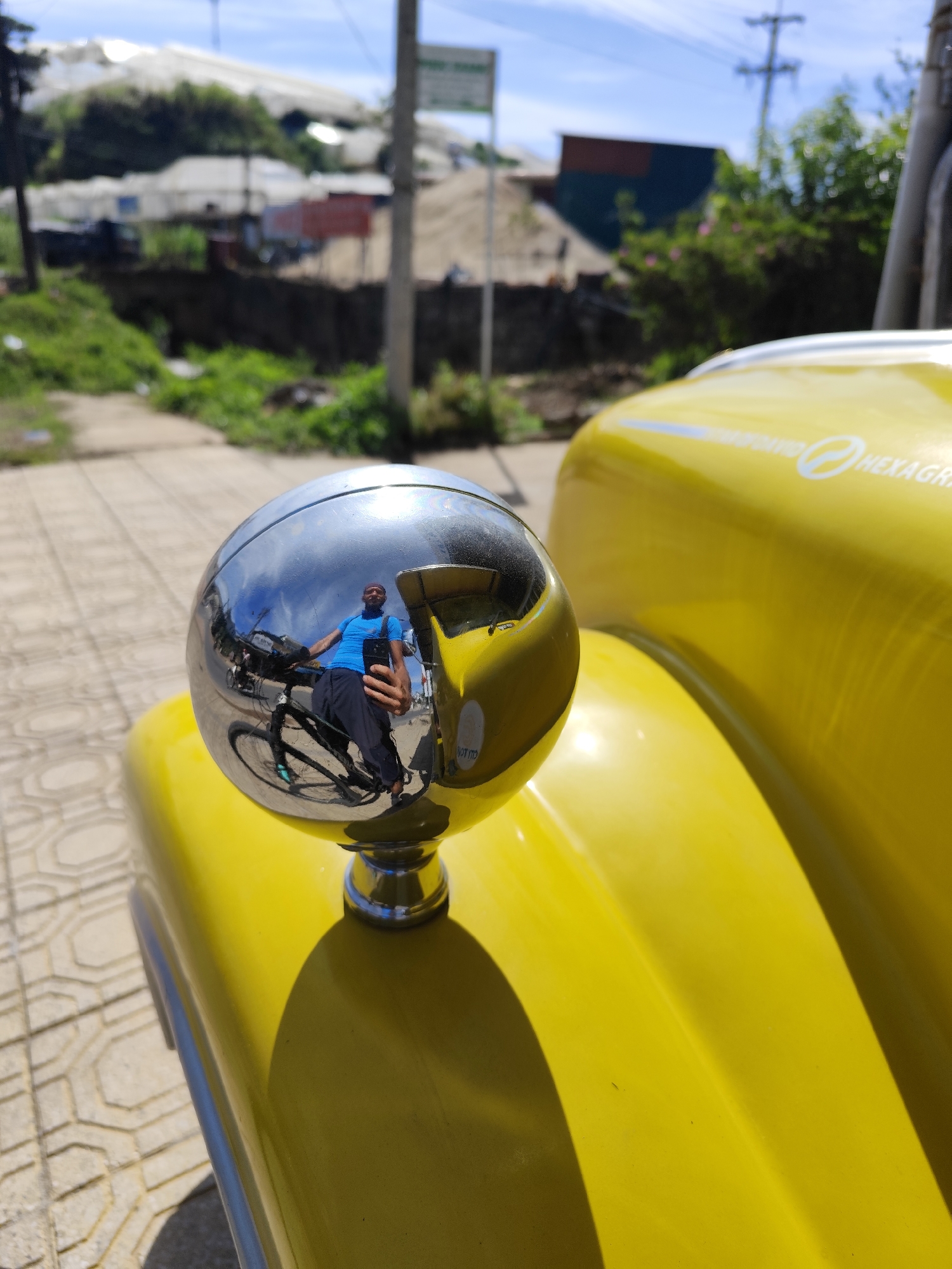 The photo shows a close-up of a shiny, spherical mirror mounted on a bright yellow vehicle, likely a type of taxi or tour vehicle, sitting on a patterned concrete surface. Reflected in the mirror is a man wearing a blue shirt and dark pants, holding a cell phone and seated on a bicycle. The background, visible both through the mirror's reflection and behind the vehicle, includes a street scene with greenery, buildings, and what appears to be agricultural structures, possibly greenhouses. In the distance, there are utility poles and other infrastructure, suggesting a semi-urban or rural location. The image was taken in Xuân Trường - Đà Lạt, Vietnam.