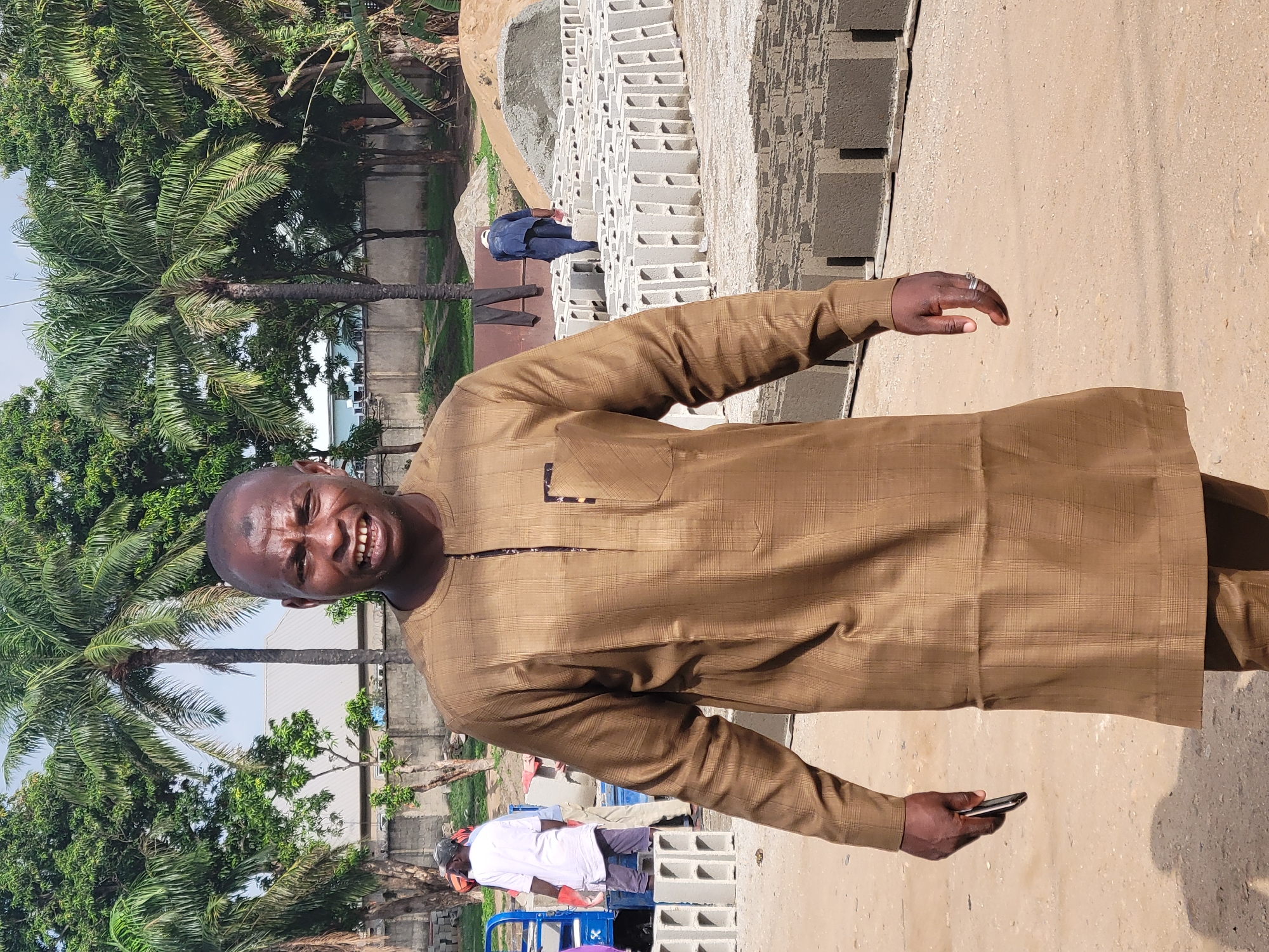 A man in a long-sleeved, brown, plaid-patterned shirt is standing in Kwamba, Nigeria, holding a smartphone. Palm trees are visible in the background. A pile of gray concrete blocks is to the side, near a concrete walkway. Another person, dressed in blue, is visible walking on the concrete blocks. A person wearing a white shirt and a face mask is seen in the background, near a blue cart filled with gray concrete blocks. The ground is light brown.