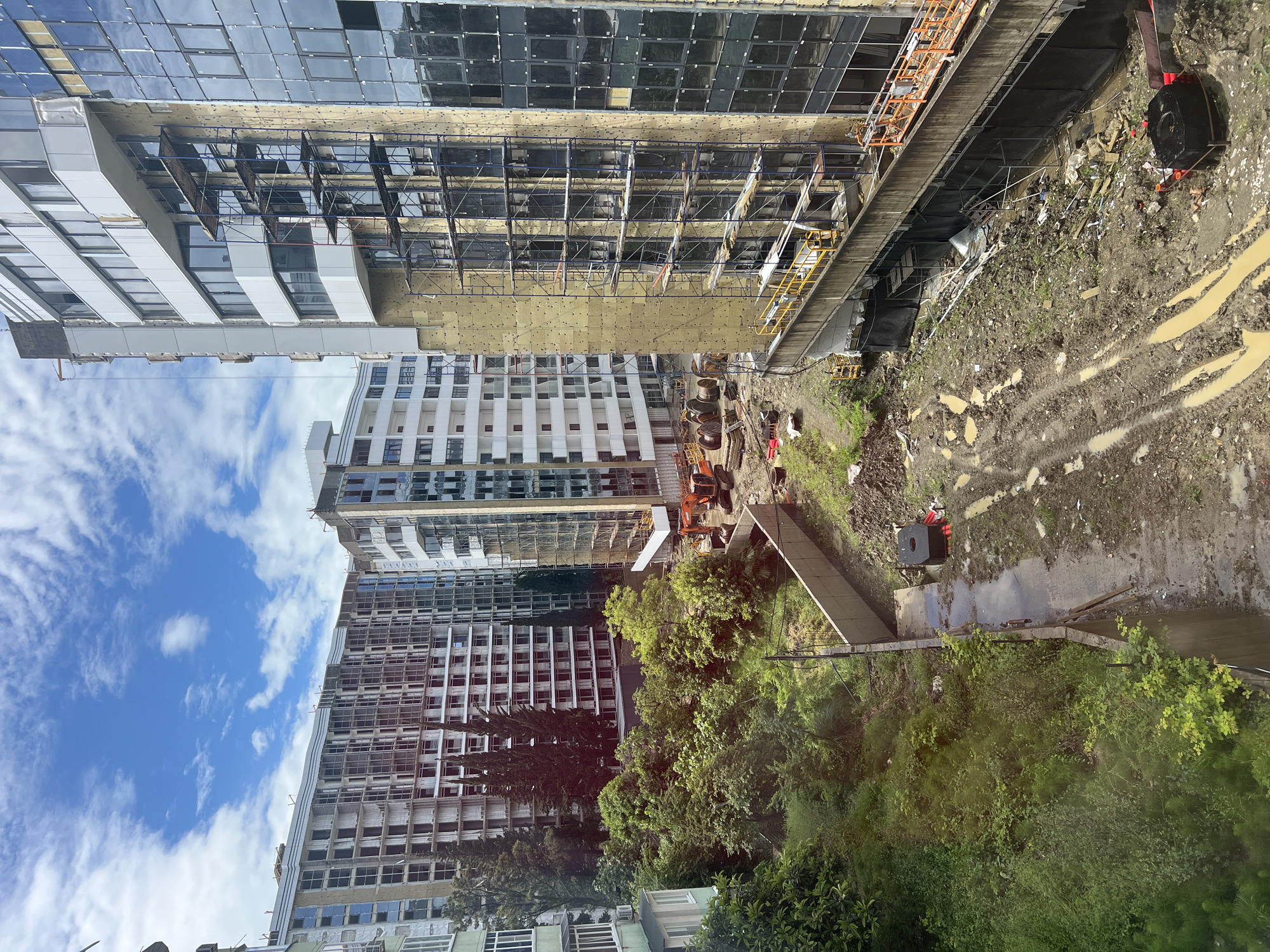 The image shows a view of a construction site adjacent to multi-story buildings, likely an urban setting.
**Buildings:** Several high-rise buildings are visible, some under construction. Scaffolding is present on one building. The architecture is modern.
**Construction Site:** The site appears to be muddy with tire tracks, indicating recent construction activity. Construction equipment is visible. There is a concrete walkway or bridge extending into the muddy area.
**Environment:** Trees and vegetation are visible near the buildings and construction site. The sky is partially cloudy with blue patches.
**Location (Implied):** This image depicts Sochi, Russia, likely in the vicinity of building construction.