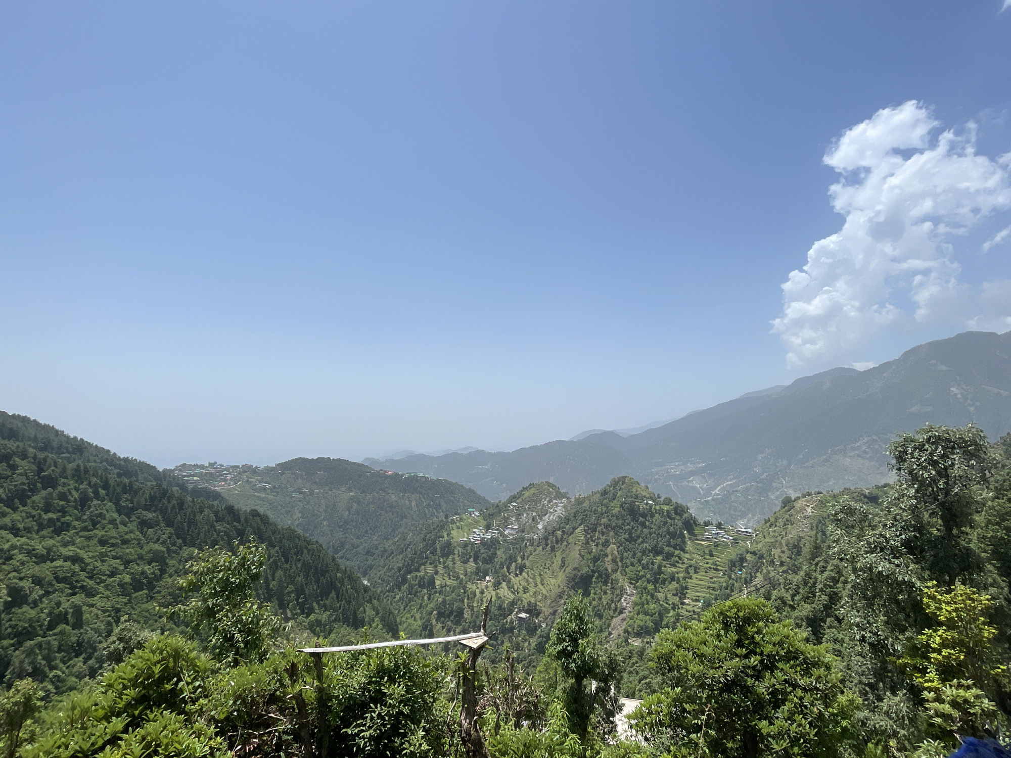 The image captures a panoramic vista of the mountainous terrain in Dharamkot, India, under a clear, blue sky with a few white clouds. Multiple verdant hills and valleys stretch into the distance, dotted with settlements consisting of small buildings. The scene suggests a serene, rural environment. In the foreground, there are trees and shrubs, adding depth to the image.