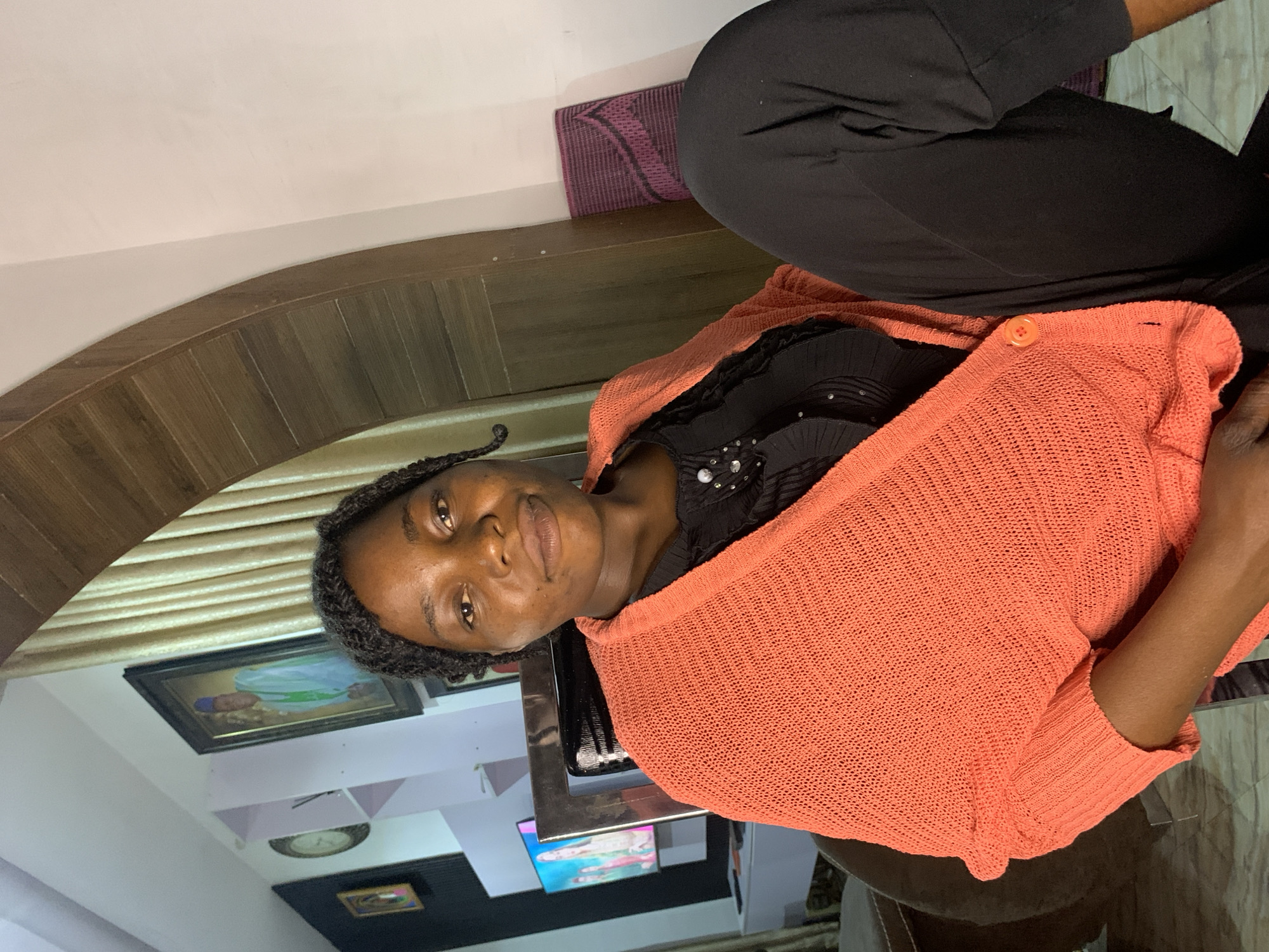 A medium shot captures a young Black woman from the chest up, seated indoors and facing the camera. She has braided hair with one strand pulled to the side. She wears a black top embellished with small shiny sequins and a vibrant coral-colored knitted cardigan open at the front, fastened with a single orange button. Her left leg is crossed over her right, and she is wearing black pants.
The background features an interior scene. To the left, a shelf unit holds framed pictures, one depicting a person in light blue and green attire, and a round clock. A television screen displays an image of people. Behind her, a textured wooden archway is visible, with a light-colored wall on the right. A rolled-up mat, likely for exercise or prayer, is propped against the wall to the right. The flooring appears to be tiled. The overall lighting suggests an indoor setting with artificial light. The location is specified as the city of Jos, Nigeria.