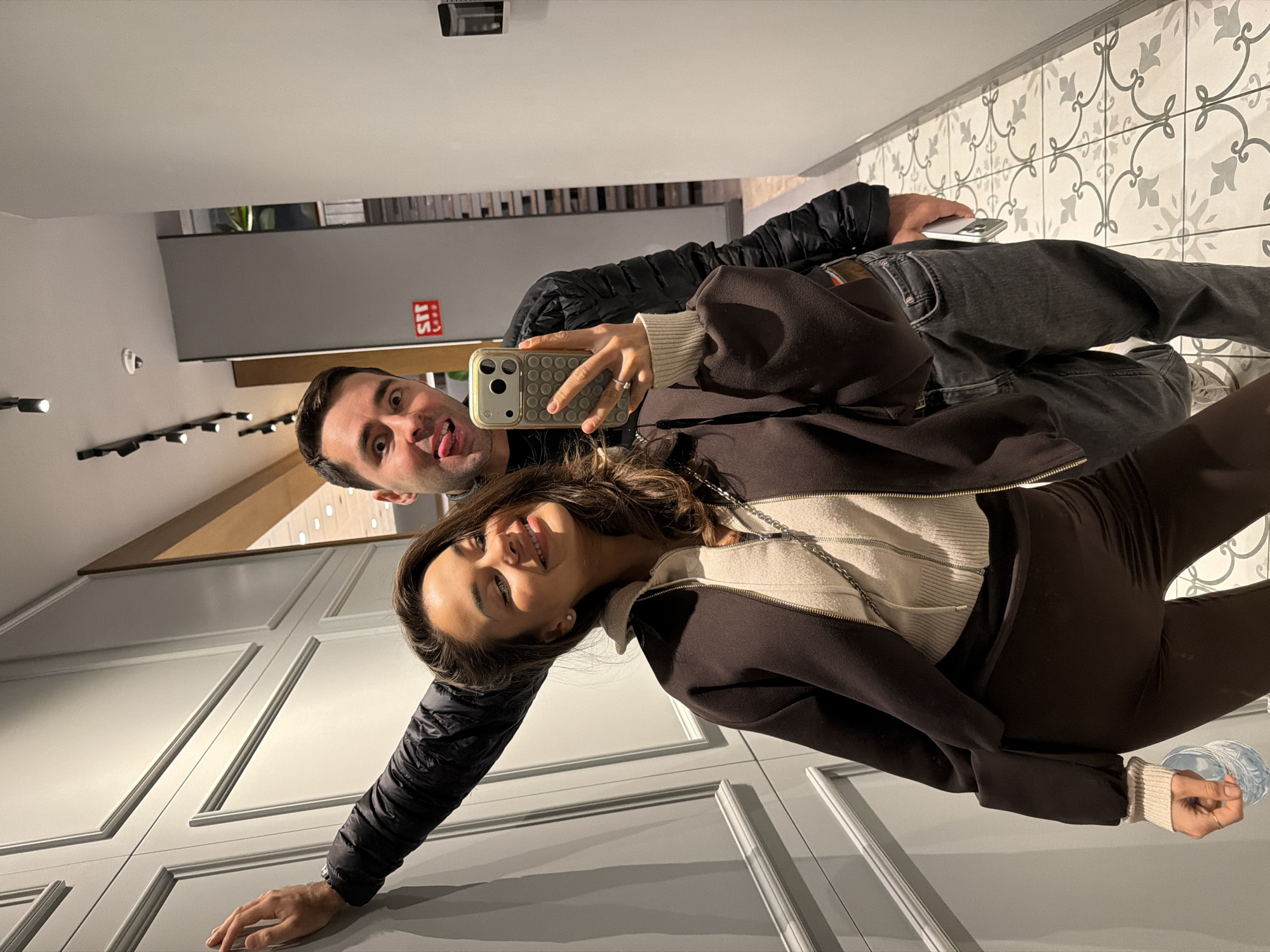 A man and a woman pose for a selfie in what appears to be an indoor location. The man, wearing a black jacket and dark jeans, is sticking his tongue out playfully towards the camera. He is holding a silver smartphone. The woman, wearing a brown zip-up jacket and a white sweater with brown leggings, smiles towards the camera and holds up a smartphone with a tan and brown case. The background reveals a modern interior design with gray paneled walls, decorative wall trim and white and gray tile flooring. In the upper left, track lighting fixtures can be seen. The location may be a commercial or residential space with decorative design elements. While there are no specific indicators in the image confirming the location as Batumi, Georgia, the interior design style is modern with decorative features that could be found in such a location.