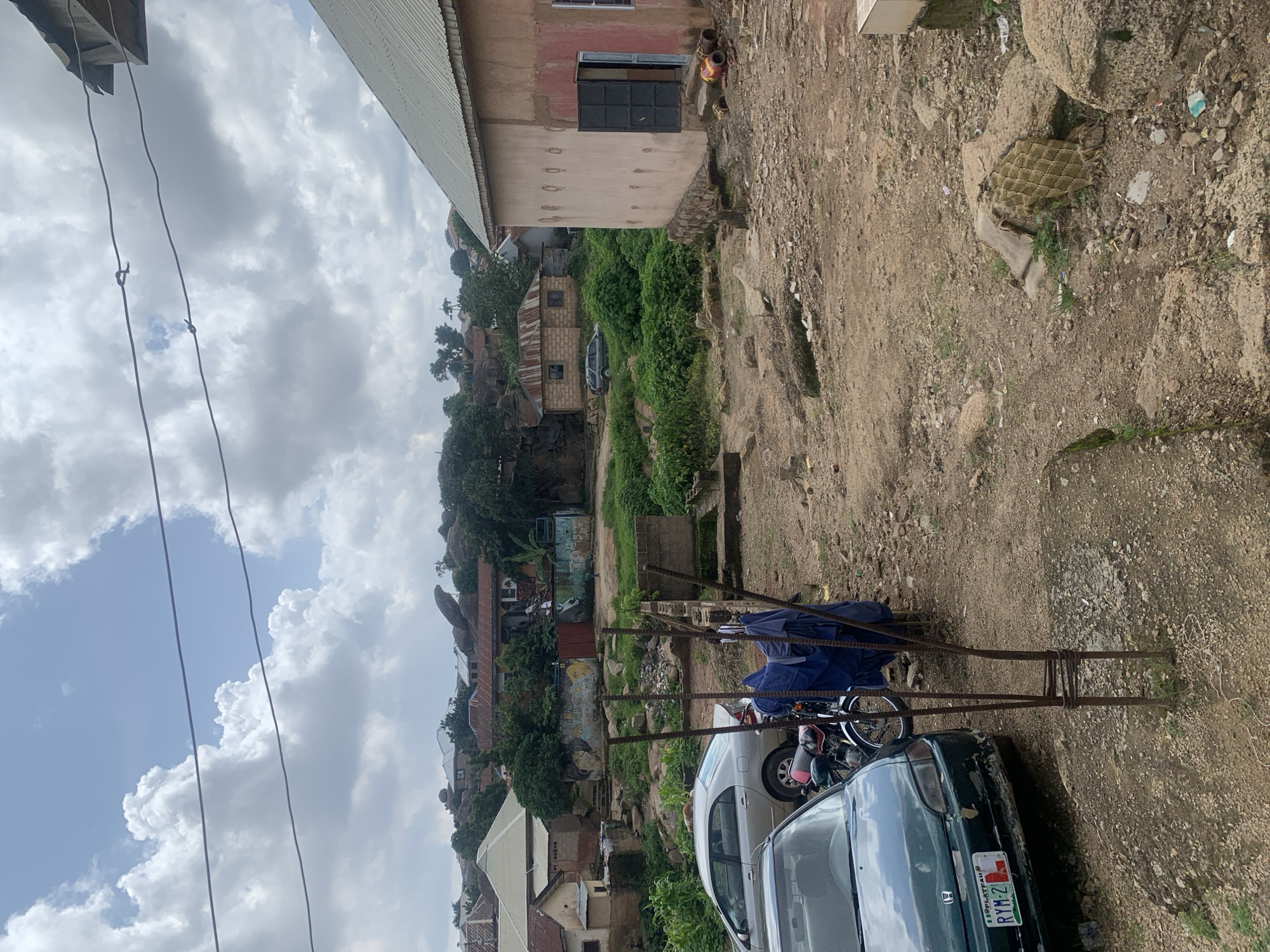 The image depicts an outdoor scene in Jos, Nigeria, likely a residential area with a hilly landscape. A dirt and gravel yard is in the foreground, with some sparse vegetation and scattered debris. Two cars are visible: a dark green sedan in the lower left corner with a license plate reading "RYM-2 L" and a silver sedan parked further back in the middle ground. A motorcycle is parked next to the green car. A rustic metal structure, possibly a part of a fence or support, stands prominently in the middle of the yard. In the background, there are several buildings with corrugated metal roofs and some with painted facades, nestled among trees and rocky outcrops characteristic of Jos. Laundry is seen hanging on a line in the middle ground. The sky is overcast with dramatic clouds. The overall impression is of a somewhat unkempt but lived-in urban environment.