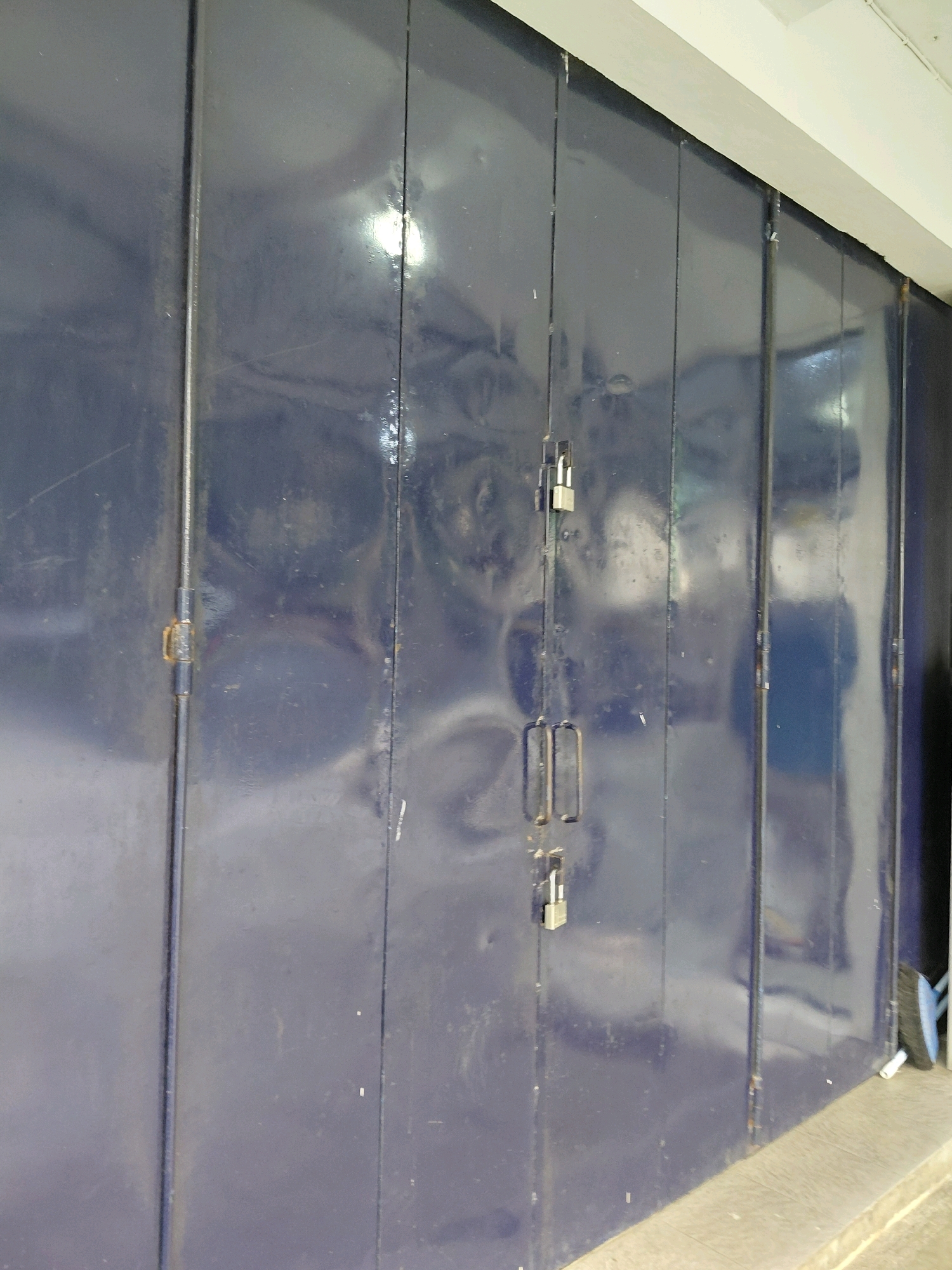 The image shows a close-up view of a closed, dark blue metal gate or rolling door. The door is segmented into several vertical panels, with visible seams between them. There are two padlocks attached to the door, one near the top and another lower down, securing it shut. The metal surface appears worn and scratched, with some reflections visible, indicating it might be painted or coated. A concrete ledge or sidewalk is visible at the bottom of the image, with a portion of a broom and a white object resting on it to the right. The scene is likely an outdoor setting, possibly in an urban or commercial area given the nature of the gate. No people are visible in the frame. The location is identified as Karanggede, Indonesia.