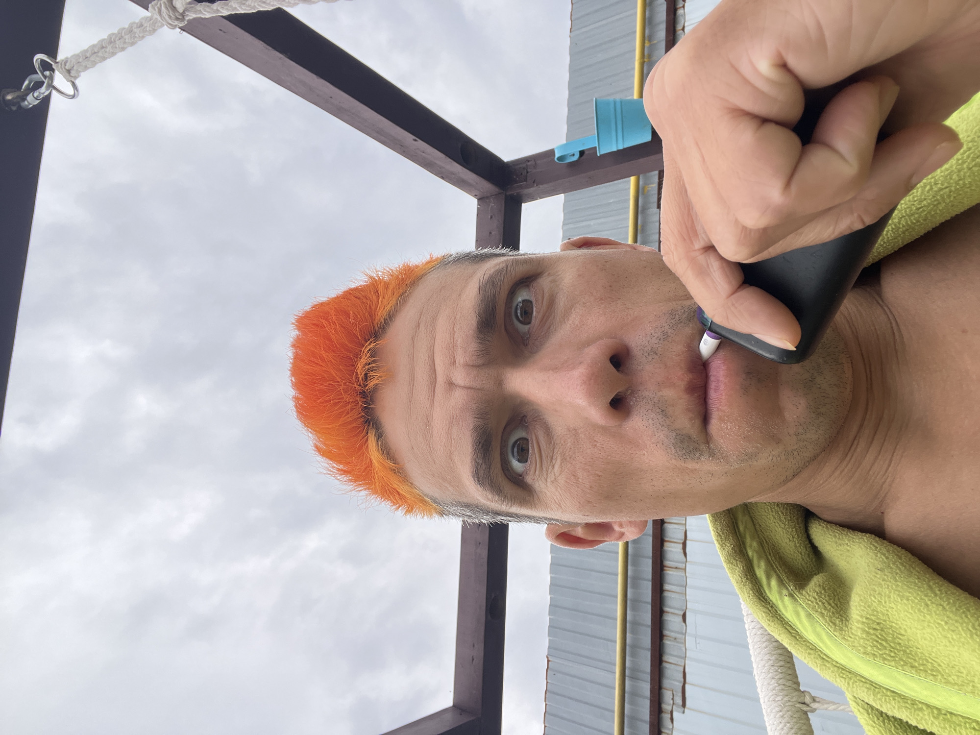 A close-up shot shows a man holding a black object to his face, potentially shaving his upper lip. He has orange hair on the top of his head with graying hair on the sides. A green towel is draped around his neck. The background includes part of a metal or wooden structure with white rope. The sky is overcast. The image appears to be taken outdoors and rotated, suggesting it may have been a selfie. There is no information available to confirm that it was taken in the city of Chaikovsky, Russia.