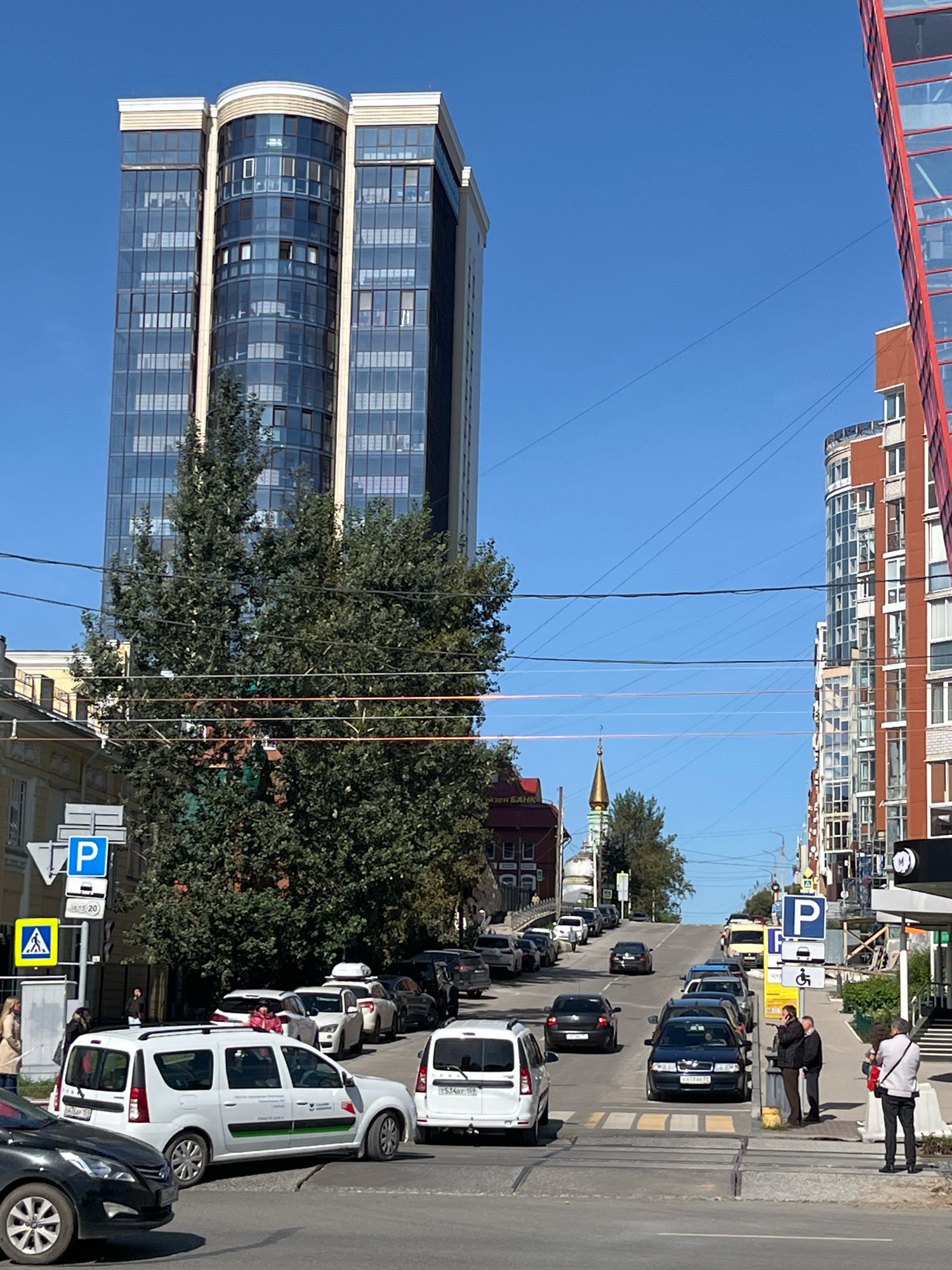 The image depicts a street scene in Perm, Russia, likely on a clear, sunny day. The foreground shows a road with several cars, including a white minivan and a black sedan in the process of crossing an intersection. A pedestrian crossing is visible. To the left, a tall, modern, multi-story building with a rounded glass facade dominates the skyline. Trees line the street, partially obscuring older, lower-rise buildings. On the right, a colorful, modern apartment building with a red facade stands. Traffic signs, including parking and pedestrian crossing symbols, are present. In the middle distance, more vehicles are visible on the uphill street, and a yellow public transportation bus is parked on the right side of the road. Further up the street, a small, golden-domed building, possibly a church, is visible. People are seen walking on the sidewalks and near the cars. The overall impression is of an urban environment with a mix of old and new architecture.