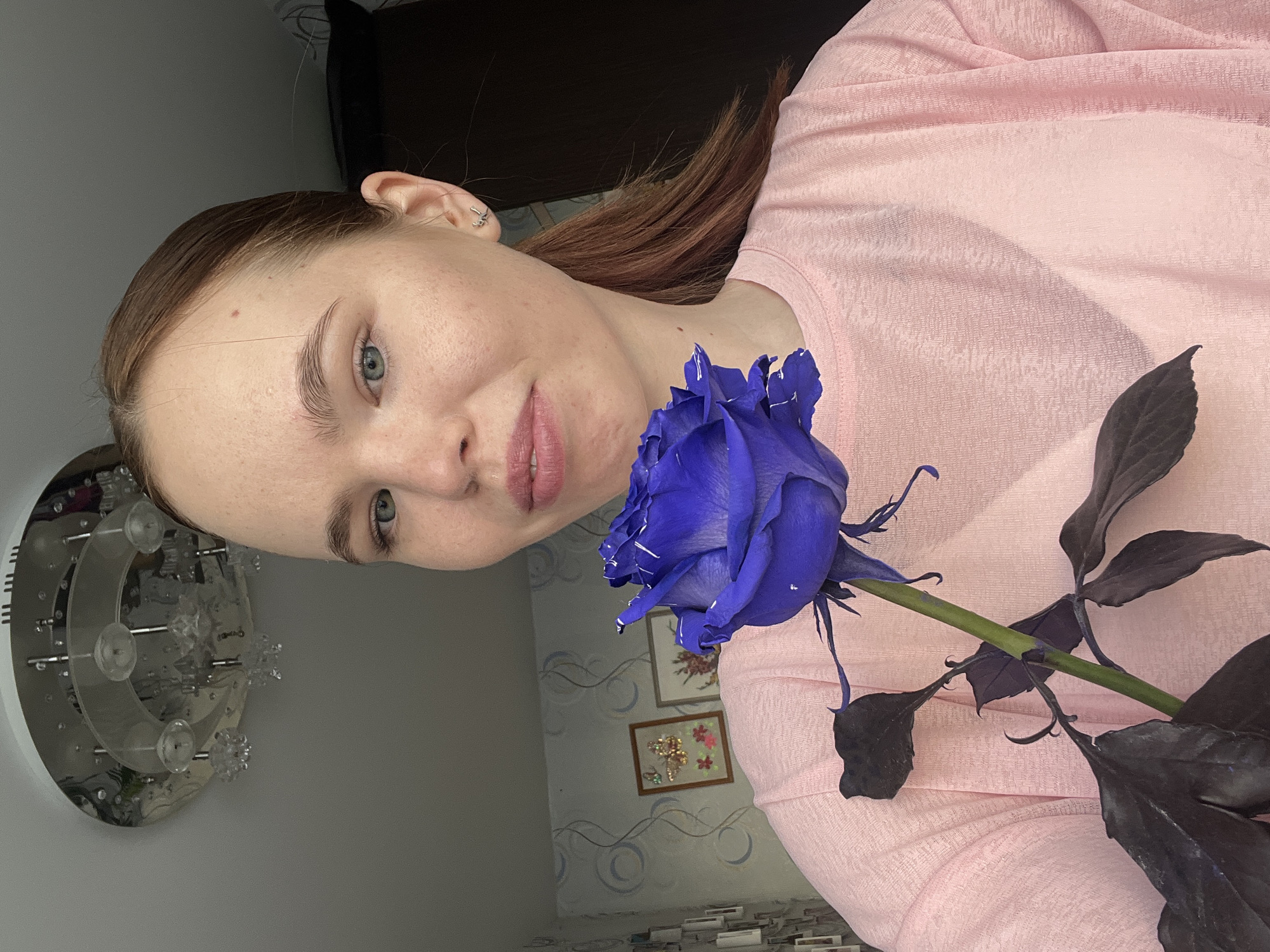 A woman with fair skin and light brown hair is holding a blue rose. She is wearing a light pink top. The background includes a wall with a decorative pattern and framed pictures. There is a light fixture visible on the left side of the image. The photograph appears to be taken indoors. It is not possible to determine the location as Chaikovsky, Russia with the provided information.