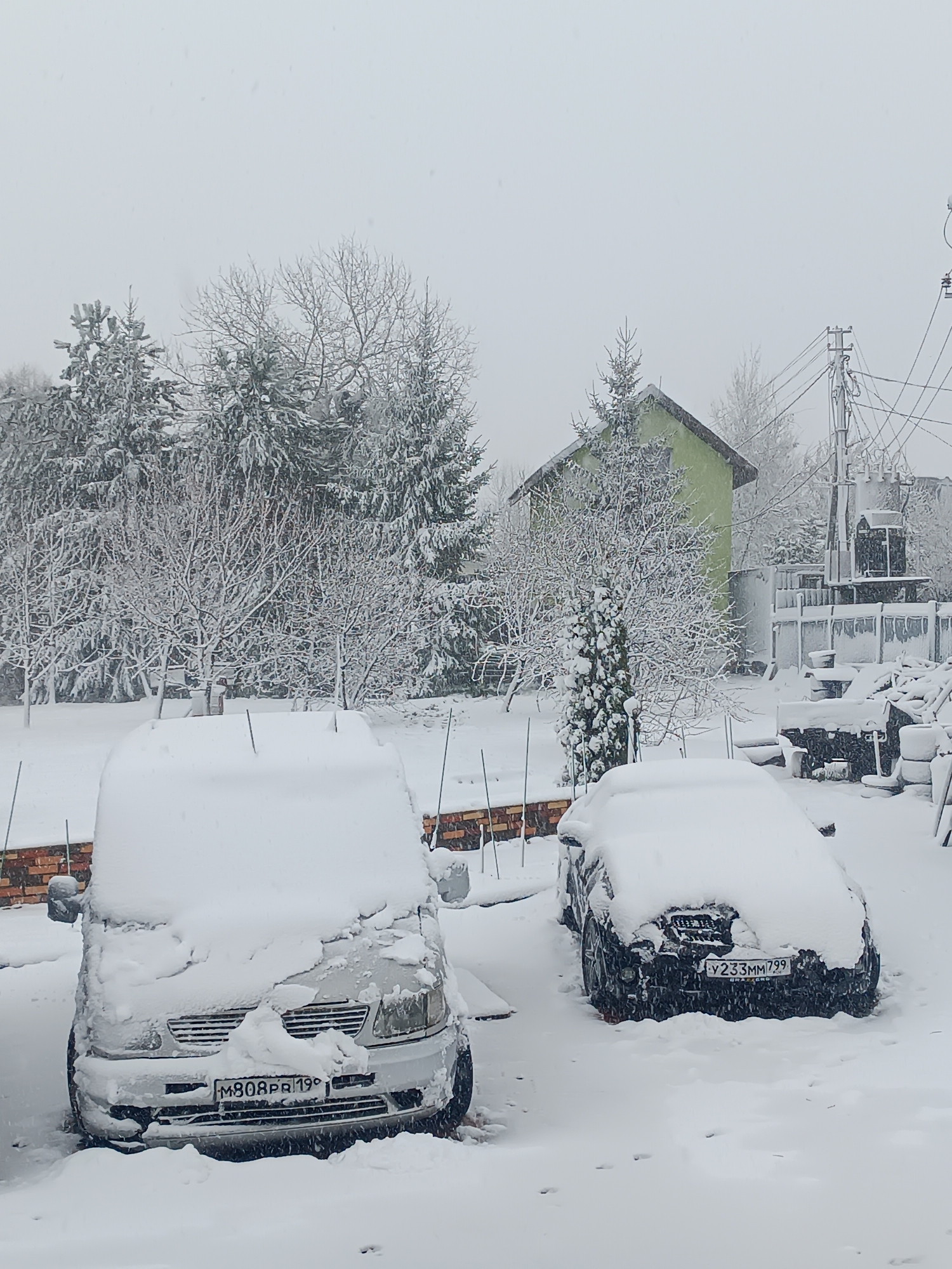 The image depicts a snowy scene in what is presumed to be Motyakovo, Russia. Two cars are heavily covered in snow in what appears to be a residential yard. The vehicle on the left is a light-colored van with the license plate "M808PA 199". The vehicle on the right is a dark-colored car with the license plate "V233MM 799". Snow blankets the ground, trees, and a green house in the background. Power lines and a utility pole are visible. There is a brick structure and some metal poles present. The weather condition is heavy snowfall.