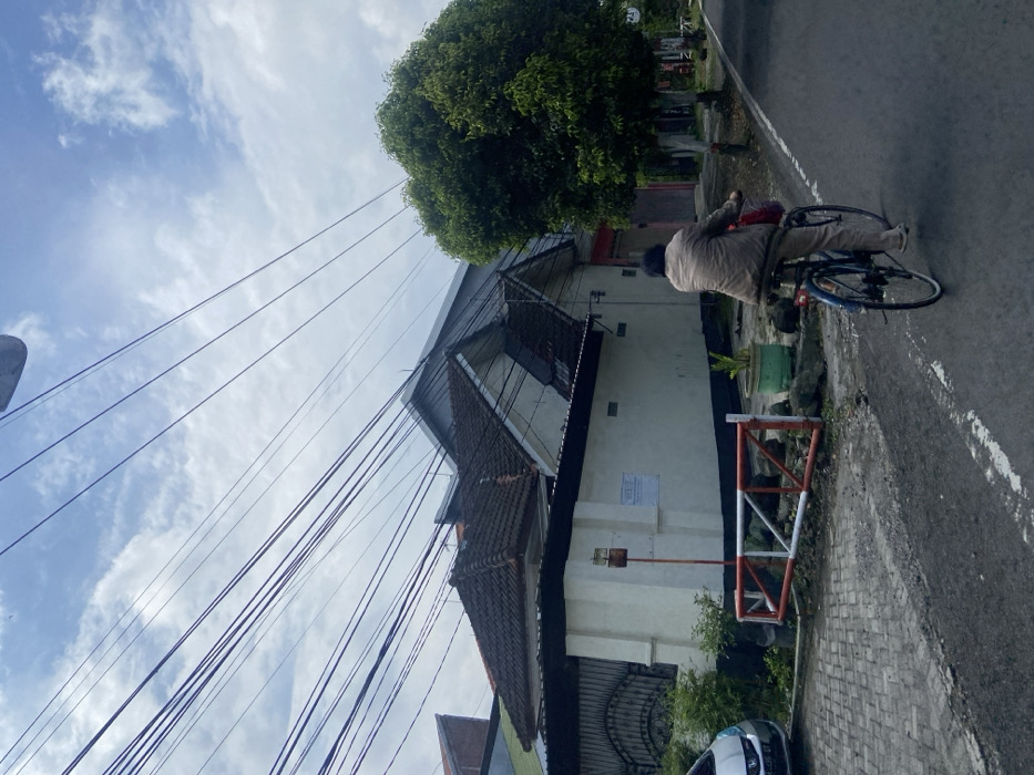The image shows a residential street in Mojokerto, Indonesia, at the location of latitude: -7.475, longitude: 112.441. A person is riding a bicycle down the street. The person is wearing a brown jacket and a red bag is attached to the front basket of the bike. There are several homes to the left of the street, and a tree to the right. There is a white car parked to the left of the frame. Many power lines stretch across the sky. There are other homes visible in the distance. The sky is cloudy and blue.