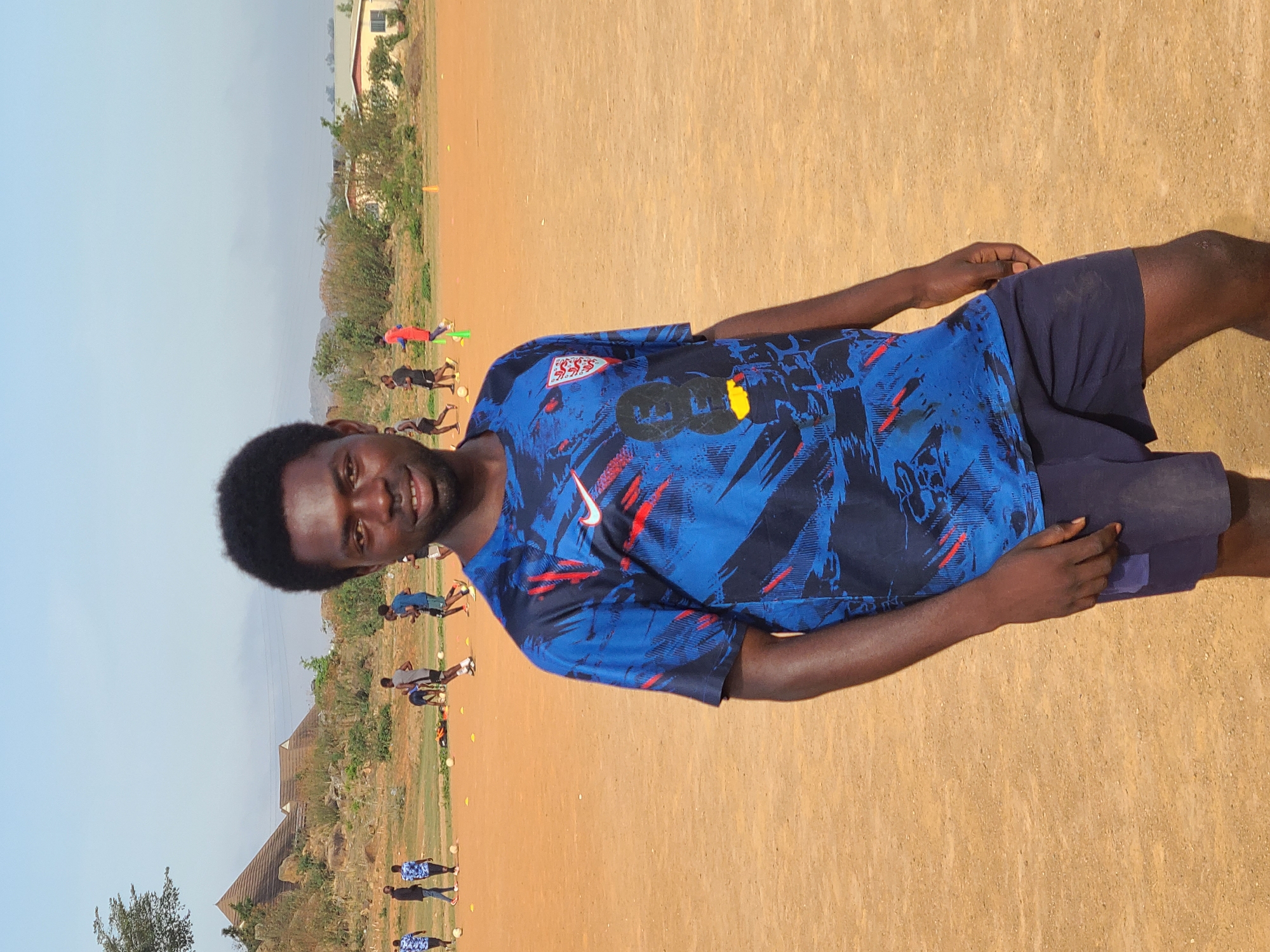 A man stands on a dirt field in what is most likely Barikin Jirga Summa, Nigeria. He wears a blue shirt with red and black accents, paired with dark blue shorts. Several other individuals are visible in the background, engaging in what appears to be soccer drills, marked by yellow cones and soccer balls on the ground. The background also includes buildings and trees. The sky is light blue.