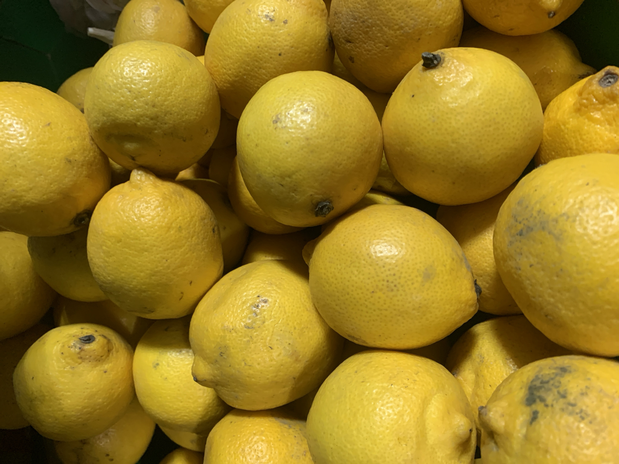 A pile of bright yellow lemons is displayed in a close-up shot. The lemons are round and have a textured rind. Some of the lemons have small blemishes or dark spots on their skins. The lighting is bright, casting shadows and highlighting the contours of the fruit. The background is dark and out of focus, suggesting the lemons are in a bin or crate.