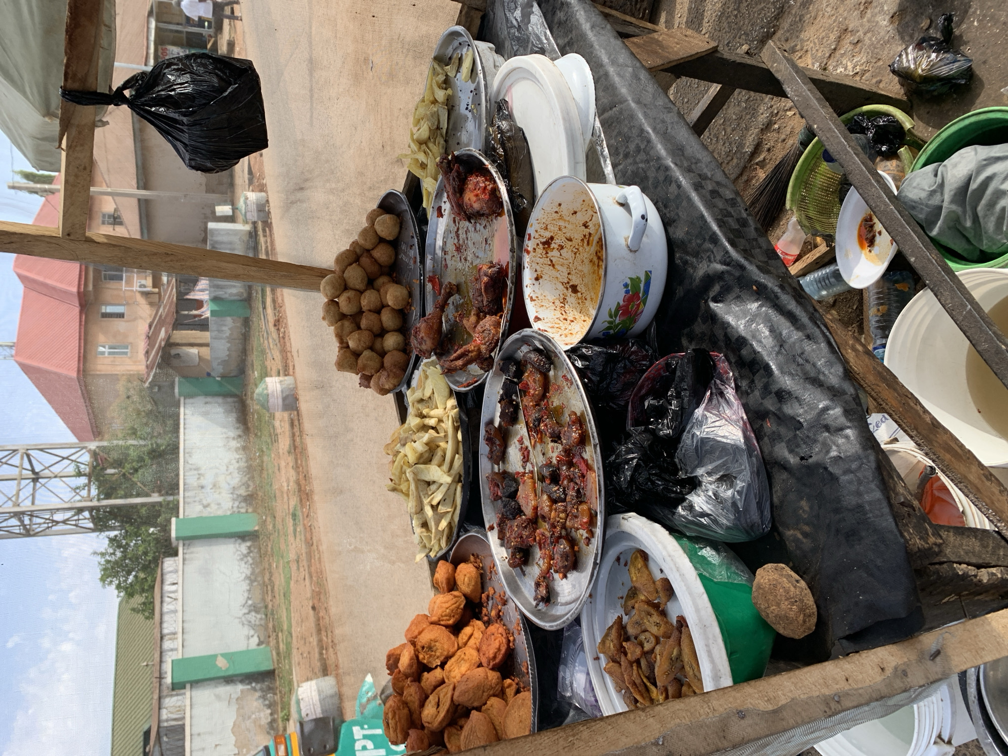 A street food vendor's makeshift stall is set up near New Stadium, Nigeria. The stall is a dark, wooden platform covered with a black tarp. Various metal platters and plastic containers hold an assortment of fried foods, including small round balls, chicken pieces, fried yam, and fried plantains. Some containers are empty or have food remnants. A stained, white enamel pot sits among the food containers. Black plastic bags are tied to the stall and strewn among the food. Discarded plastic bottles and food containers are visible near the stall. In the background, a fence and buildings are visible under a partly cloudy sky.