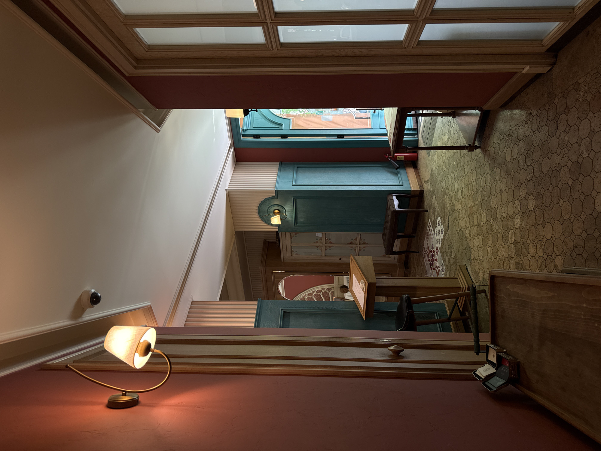 Captured in Yerevan, Armenia, the image depicts an interior hallway. The foreground features a vintage-style table lamp with a beige shade on a reddish-brown wall, along with a security camera mounted higher on the wall. Adjacent to the wall is a staircase with wooden steps. Further down the hallway, there is an array of furniture including wooden cabinets and chairs. The flooring is patterned with a hexagonal design, while the ceiling is structured with square panels. A window in the distance provides a glimpse of the cityscape outside. The architectural style of the interior is classic with teal and white accents. No people are visible.