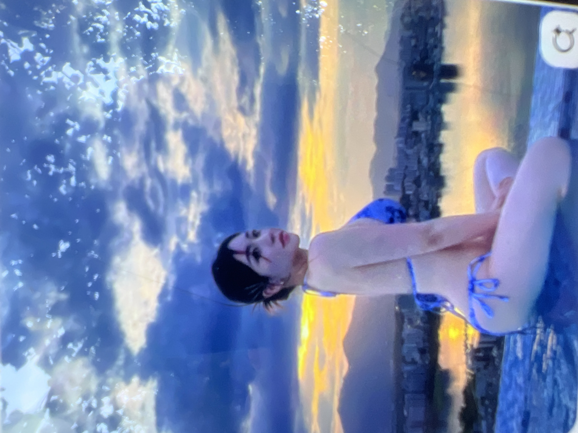 The image shows a young woman in a blue bikini sitting in an infinity pool. She is looking to her right, away from the camera, with her head tilted slightly. Her hair is dark and pulled back in a messy bun. She is wearing dark eye makeup. The background depicts a city skyline with mountains in the distance. The sky is cloudy with hues of orange and yellow from a sunset or sunrise. The water of the infinity pool reflects the sky and the city. There is a circular icon with an arrow in the bottom right corner of the image. The location is not Kara-Dıykan, Kyrgyzstan; this scene appears to be a city with a body of water and mountains, possibly a coastal or lakefront urban area.