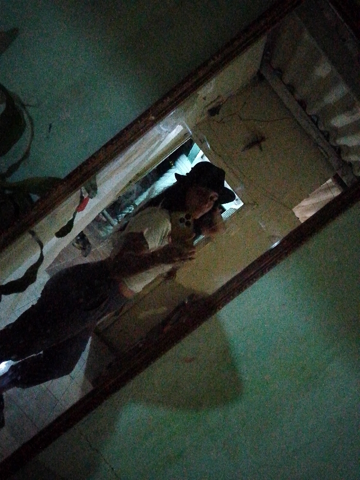 The image depicts a female individual captured in a mirror selfie within an interior space, stated to be located in Juan Griego, Venezuela. The exact position within the city is not discernible from the visual evidence.
The individual, wearing a black wide-brimmed hat, a light-colored short-sleeved top with a dark circular design on the chest, and dark-colored trousers, is holding a dark-colored smartphone in both hands to photograph her reflection. Her face is partially obscured by the phone and the hat. Her long, dark hair extends below the hat.
The mirror is rectangular, framed by a dark, possibly wooden, border. The surrounding environment outside the mirror's reflection shows a textured, teal-green wall to the right of the frame (viewer's perspective) and an indistinct dark object, possibly a plant, to the left. The reflection within the mirror reveals a lighter-colored wall or ceiling behind the individual, displaying visible cracks or markings. Below the individual's waist, a faint tiled floor or patterned surface is visible in the reflection. The scene is dimly lit, with notable image grain, suggesting low-light conditions. Bright highlights are visible in the background reflection, possibly from an external light source or window.