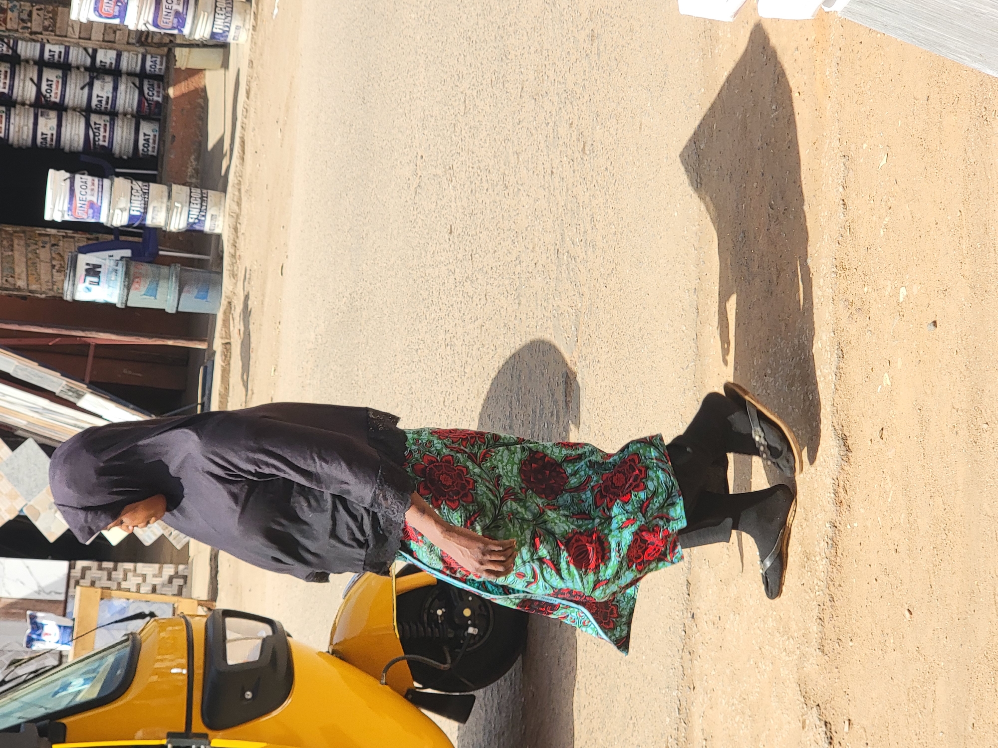 A woman wearing a black head covering, black long-sleeved shirt, and a colorful patterned skirt is sitting in a yellow vehicle. The vehicle is parked on a paved road. To the left of the vehicle, stacks of paint cans are displayed outside of a shop. The background shows a beige pavement. The scene is set in Maiburiji, Nigeria.