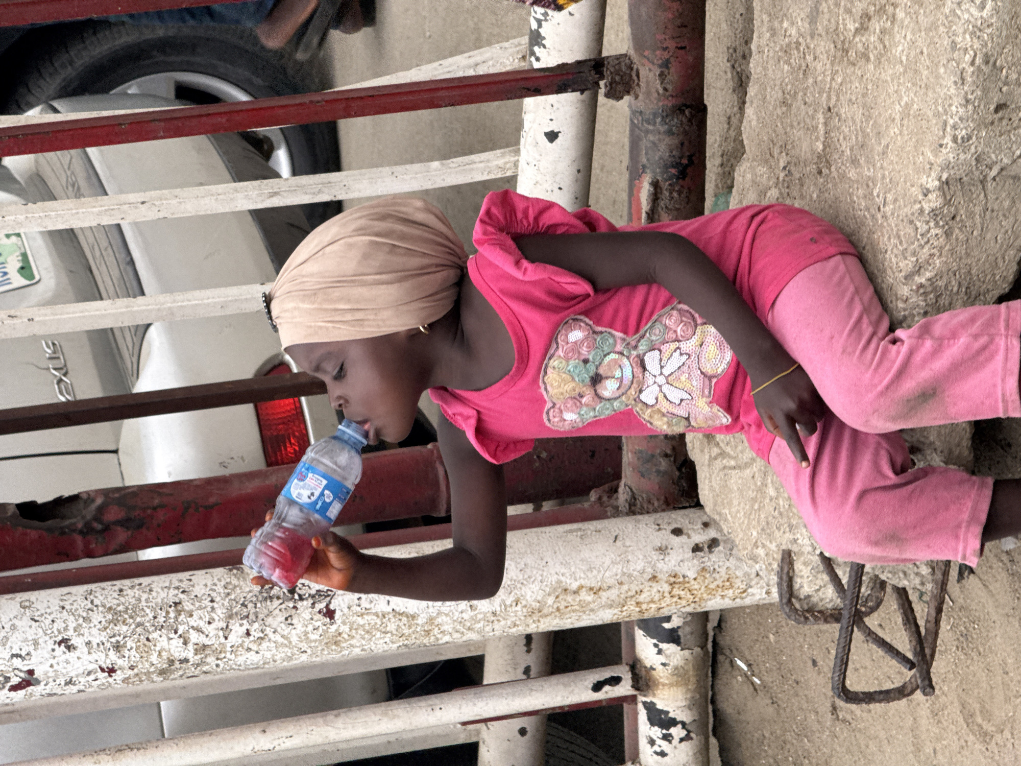 In Lekki, Nigeria, a child wearing a tan head wrap, pink shirt, and pink pants is lying on a dirty concrete surface, drinking from a clear plastic water bottle. A partially visible white car, a metal barricade with peeling paint, and a rusty metal structure are also present in the scene.