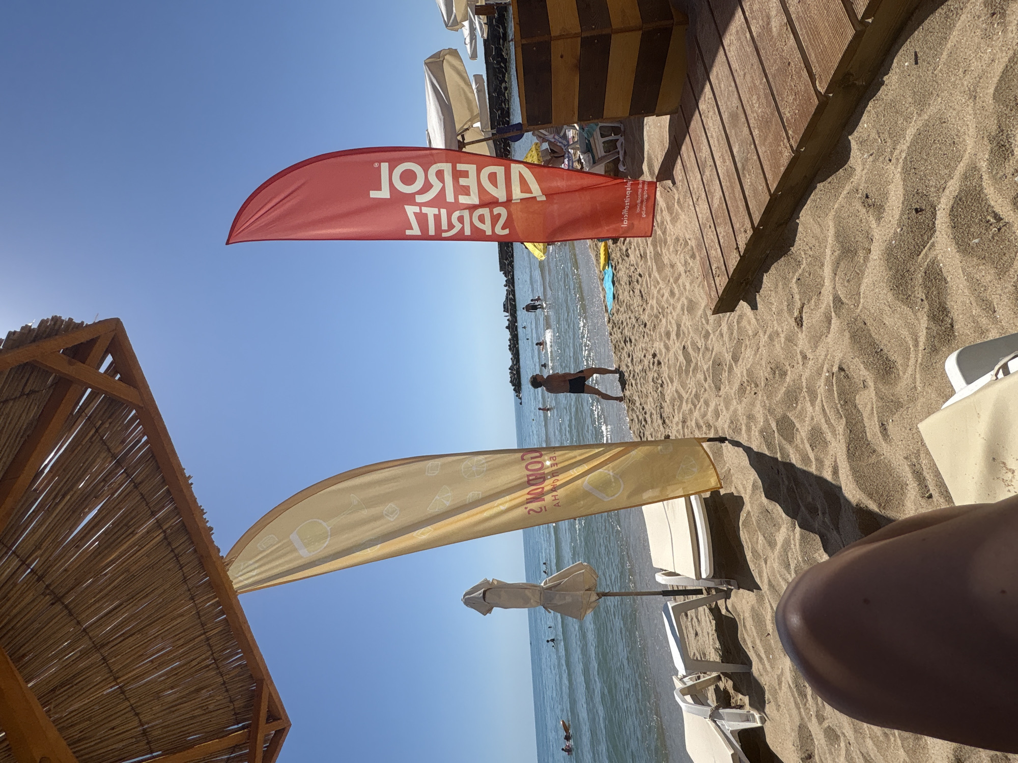 The image depicts a beach scene on a sunny day. In the foreground, a person's leg is visible, resting on a lounge chair. Behind the leg, the sand stretches towards the water, dotted with lounge chairs and umbrellas. Two large flags, one yellow and one red, are prominently displayed. The yellow flag features an image of a wine glass and ice cubes, along with text that appears to be in Cyrillic. The red flag has text in white, also in Cyrillic, and what seems to be a logo. In the background, the ocean is calm with a few people swimming or wading. A breakwater is visible in the distance, and beyond that, more beach umbrellas and structures. The sky is clear and blue. The overall atmosphere is relaxed and summery.