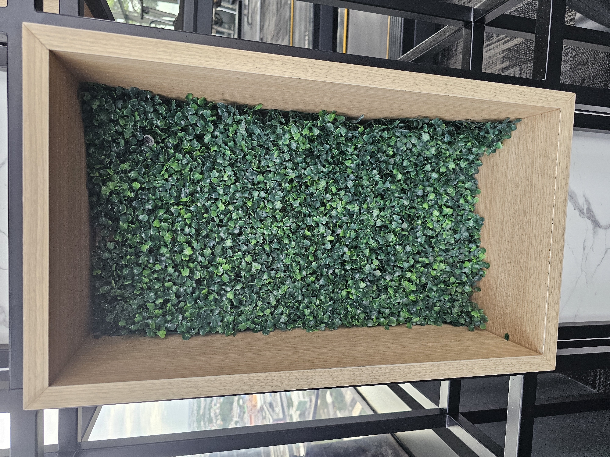 The image shows a close-up view of a wooden frame containing artificial greenery. The frame is light brown and has a minimalist design. The greenery appears to be made of small, dense, dark green leaves. The frame is part of a larger shelving unit with black metal framing. In the background, through a window, a cityscape is visible with buildings and roads, suggesting an urban environment. The lighting appears to be natural daylight.