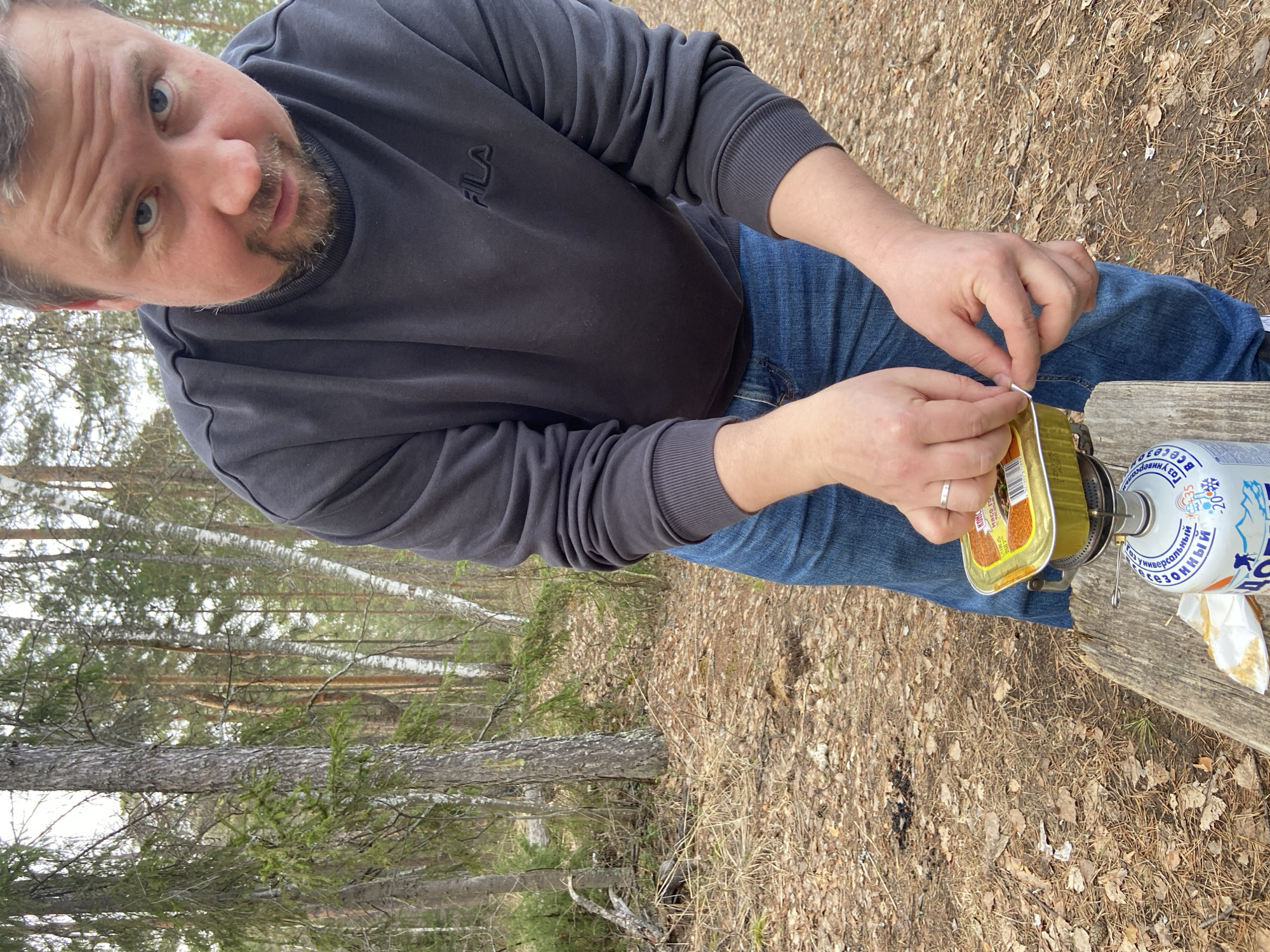 A man in a gray sweatshirt and blue jeans is seated outdoors on a wooden surface near a forest. He is opening a tin can with his hands; the can is gold-colored and appears to contain food. To the right of the can is a portable camping stove with a gas canister attached. The ground is covered with fallen leaves and foliage. The location is Chaikovsky, Russia, as indicated by the Cyrillic text on the gas canister, and the landscape featuring birch trees is typical of the region.