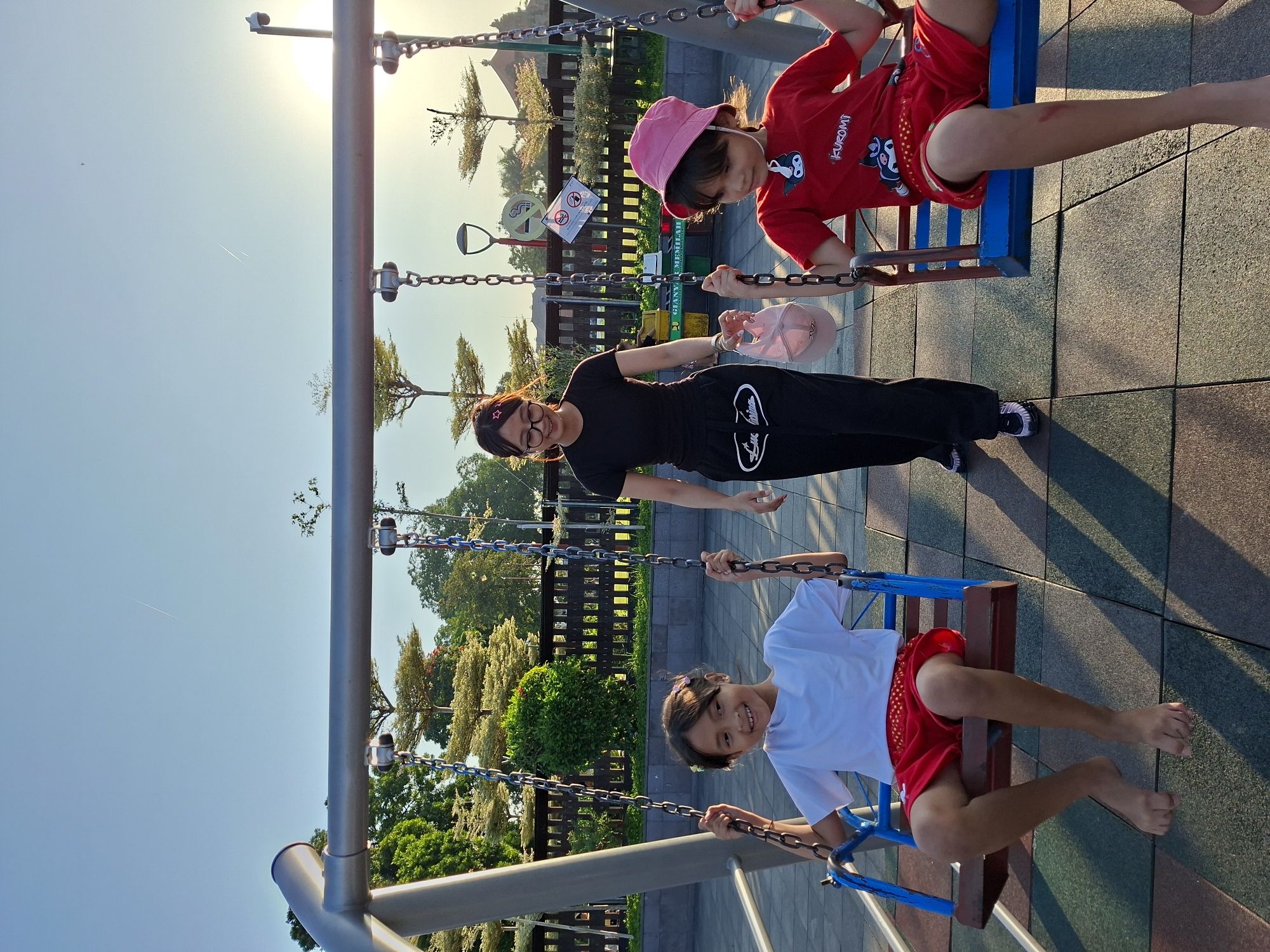 In Gianyar, Indonesia, two children are on swings at a playground. The child on the left, wearing a white t-shirt and red shorts, smiles at the camera. The child on the right, wearing a red t-shirt with cartoon characters and a pink hat, looks down. A woman in a black t-shirt and black pants stands between them, holding a pink hat. The playground has a blue and red metal swing set with chains. The ground is made of colorful rubber tiles. In the background, there are trees and a wooden fence. The sun is setting, casting a warm glow on the scene.