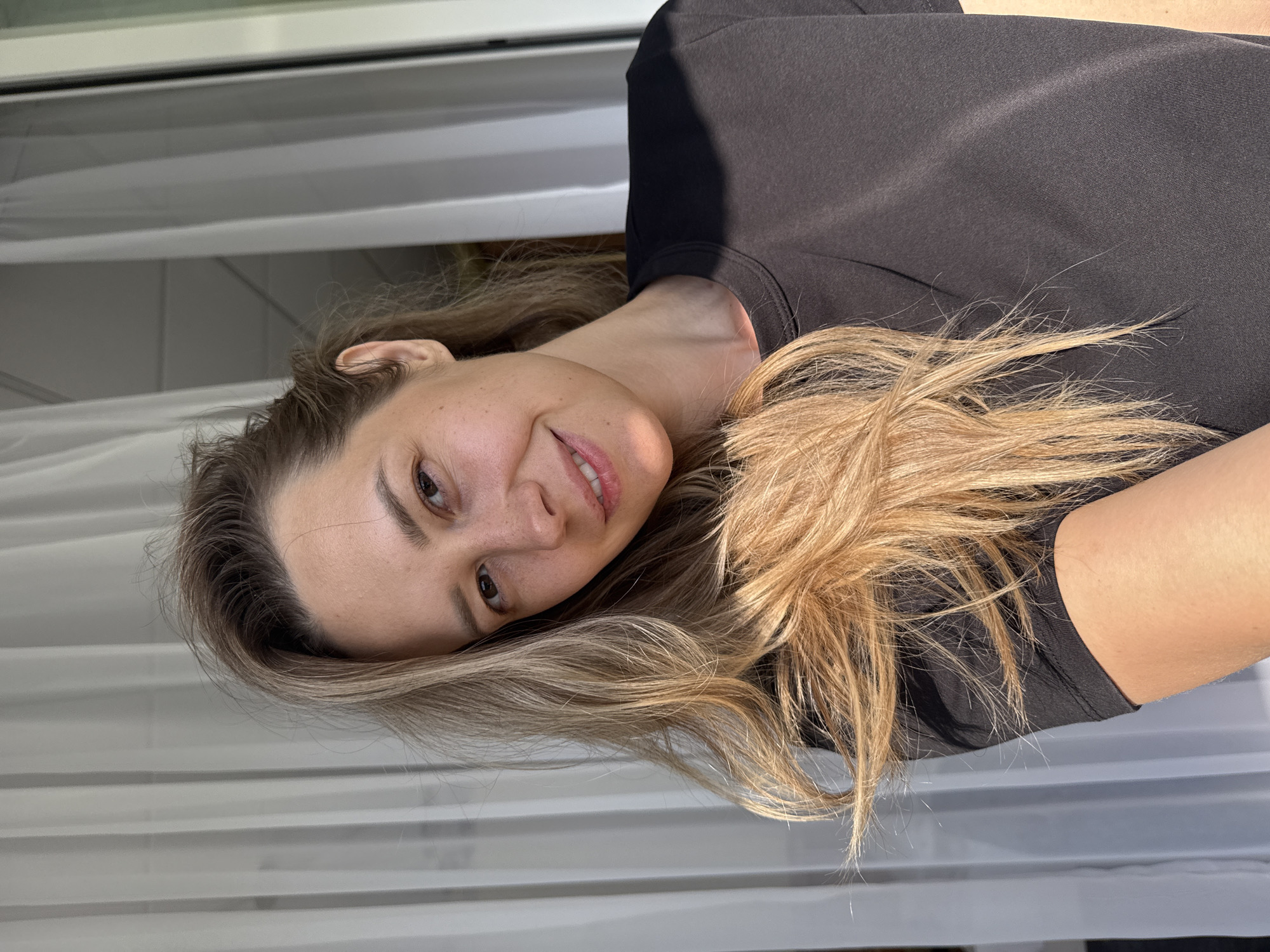 A close-up, head-and-shoulders selfie features a woman with light brown hair, wearing a black t-shirt. She is positioned in the center of the frame, with her head tilted to her right. Her eyes are looking towards the camera, and she has a subtle smile. Sunlight casts a shadow across her right shoulder. Behind her, sheer white curtains are visible, with a hint of outdoor light and a vertical window frame on the right side of the image. The background suggests an indoor setting, possibly a home or hotel room. The overall lighting is bright and natural, indicating it is daytime. The scene appears to be located in Nebug, Russia, as per the prompt, though no specific landmarks or indicators are present in the image to confirm this.