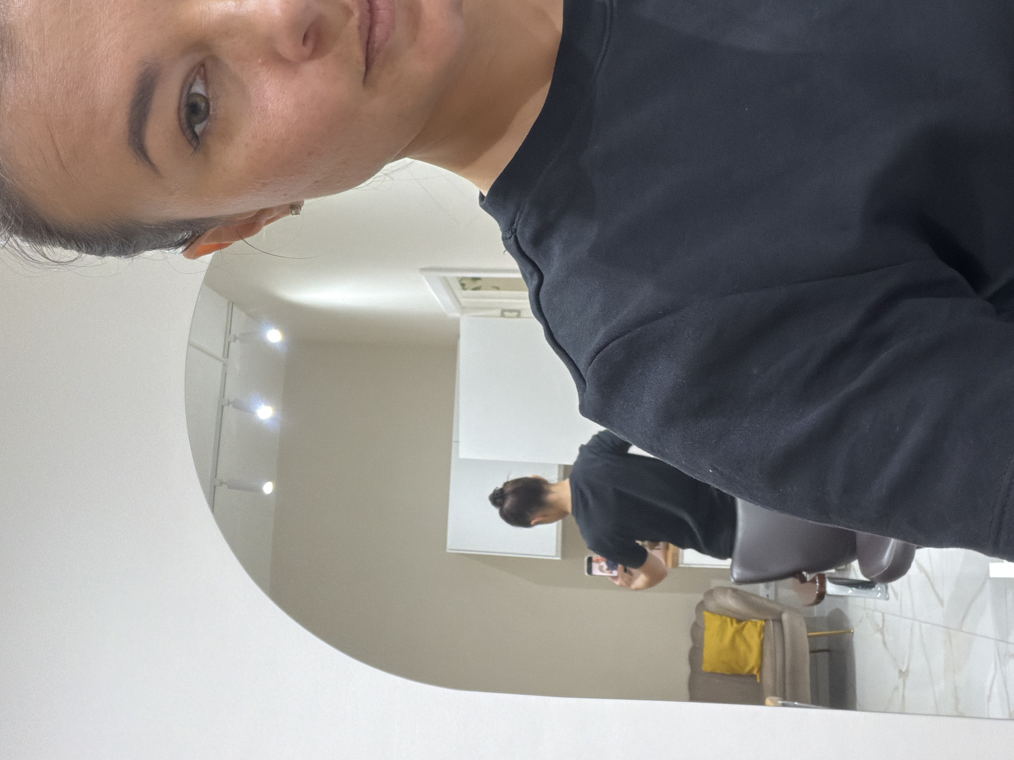 A woman is taking a selfie in front of a large, rounded mirror. The foreground is dominated by her face and the upper portion of a black sweatshirt she's wearing. The reflection in the mirror shows the woman sitting in a chair, holding a phone up to take the picture. The background shows a salon setting. The location cannot be determined from the image.