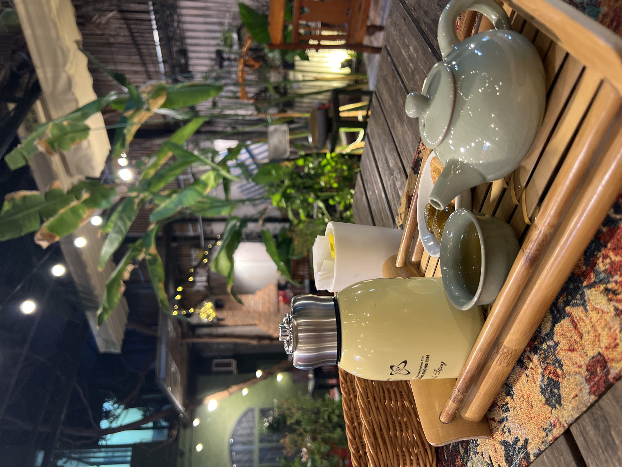 The image shows a tranquil tea setting on a wooden table, likely in an outdoor or semi-outdoor area in Nha Trang, Vietnam, based on the combination of the subject of tea service and the presence of lush tropical plants and outdoor lighting.
Subjects/Objects:
* A light blue-green ceramic teapot is positioned on a bamboo tray with a matching teacup. Tea is being poured from the pot into the teacup.
* Next to the teacup and teapot is a cylindrical, insulated drink container or thermos of pale yellow color.
* There is a small, white container holding thin, yellow items.
* The bamboo tray on which these items are arranged is lined with a patterned mat or cloth.
* The table is constructed of dark wood, and a portion of a wicker basket is visible.
* In the background, there are banana trees and other tropical plants, suggesting a garden or courtyard setting.
* Outdoor lighting, including string lights and lanterns, creates a soft, ambient atmosphere.
* Architectural elements such as corrugated roofing and a brick structure are faintly visible in the background, adding to the sense of location.
Actions/Interactions:
* The primary action is the pouring of tea from the teapot into the teacup, indicating a moment of preparation or service.
Environment:
* The setting is a relaxed, outdoor or semi-outdoor area with a tropical atmosphere, potentially a tea house, cafe, or private garden.
* The combination of the bamboo and wooden elements with lush foliage suggests a blending of natural and designed spaces.
* The lighting indicates an evening or nighttime setting, enhancing the ambiance.