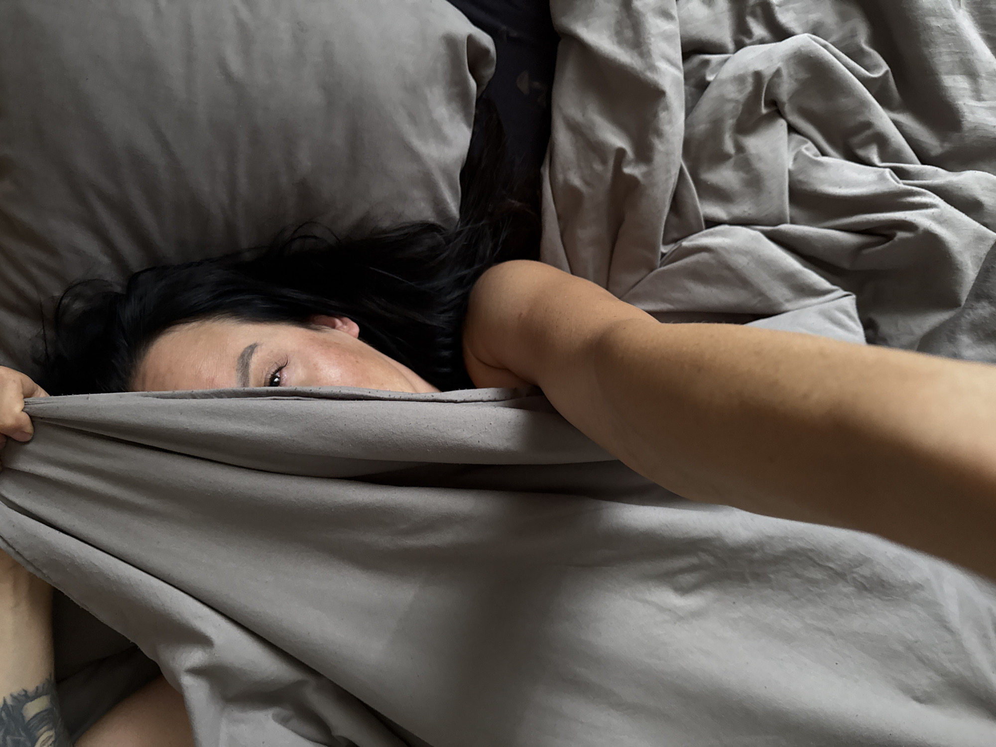 This media file shows a close-up of a person in bed. The person has dark hair and an eye visible, peeking out from under a gray blanket. Their arm is extended, also partially covered by the blanket, and a tattoo is visible on their left forearm. The setting appears to be a bedroom, with pillows and crumpled gray bedding filling the frame. The lighting suggests it could be daytime, possibly morning or evening, given the soft, diffused light. No text is visible in the image. The overall impression is one of relaxation or perhaps waking up.