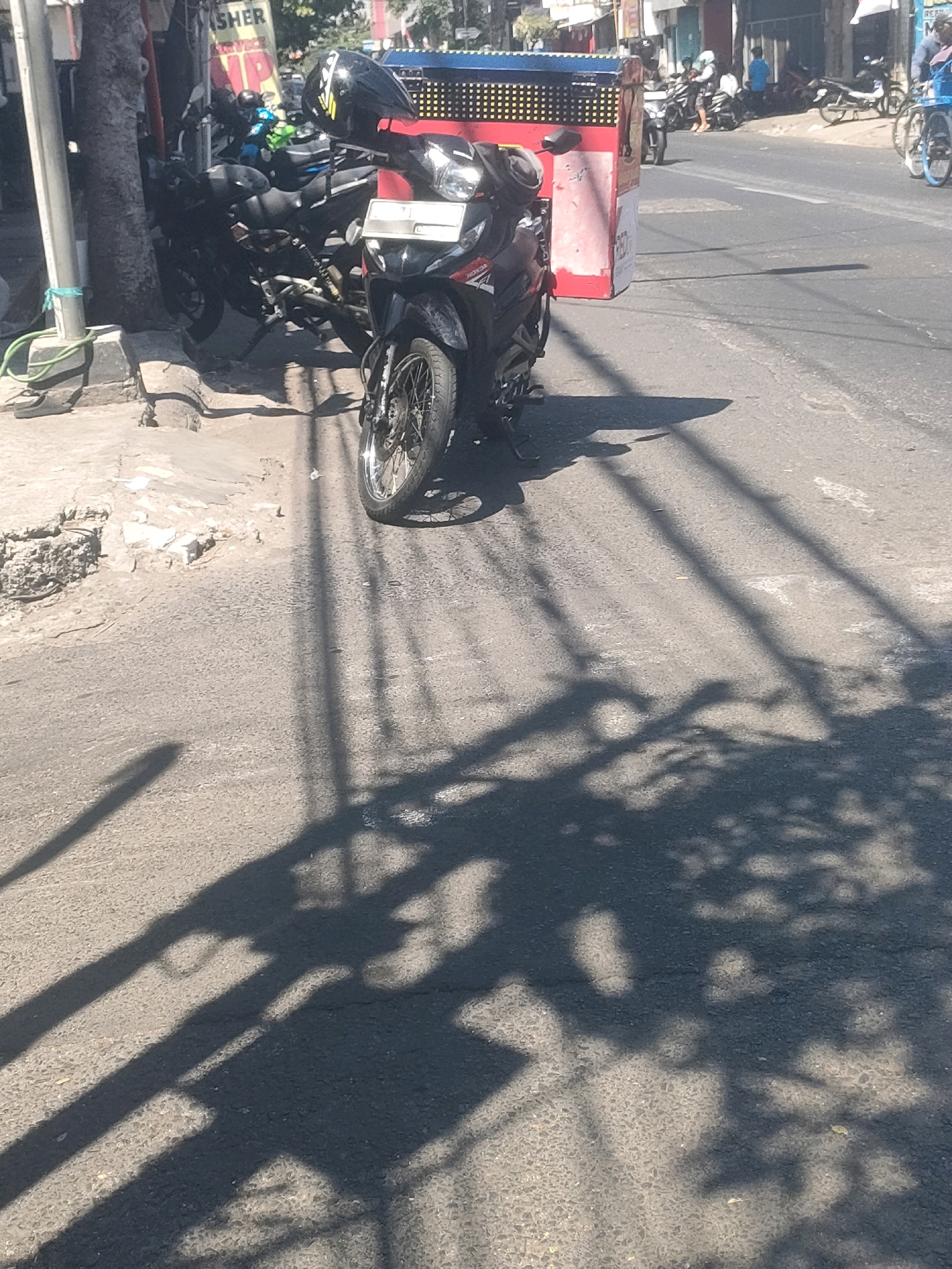 The image depicts a street scene in Surabaya, Indonesia, likely on a sunny day given the prominent shadows. The foreground features a black motorcycle parked on the asphalt road, its front wheel and part of its body visible. A helmet with yellow stripes rests on the handlebars. To the left of the motorcycle, a concrete sidewalk is visible, with a tree trunk and a green hose. Further in the background, other motorcycles are parked, some appearing to be in a line. A red and blue food cart with yellow lights is positioned behind the motorcycles. Several individuals are sparsely distributed in the background, one on a motorcycle and another walking. The street appears to be a typical urban road with buildings and other vehicles in the distance. The overall impression is a snapshot of daily life on a bustling street in Surabaya.