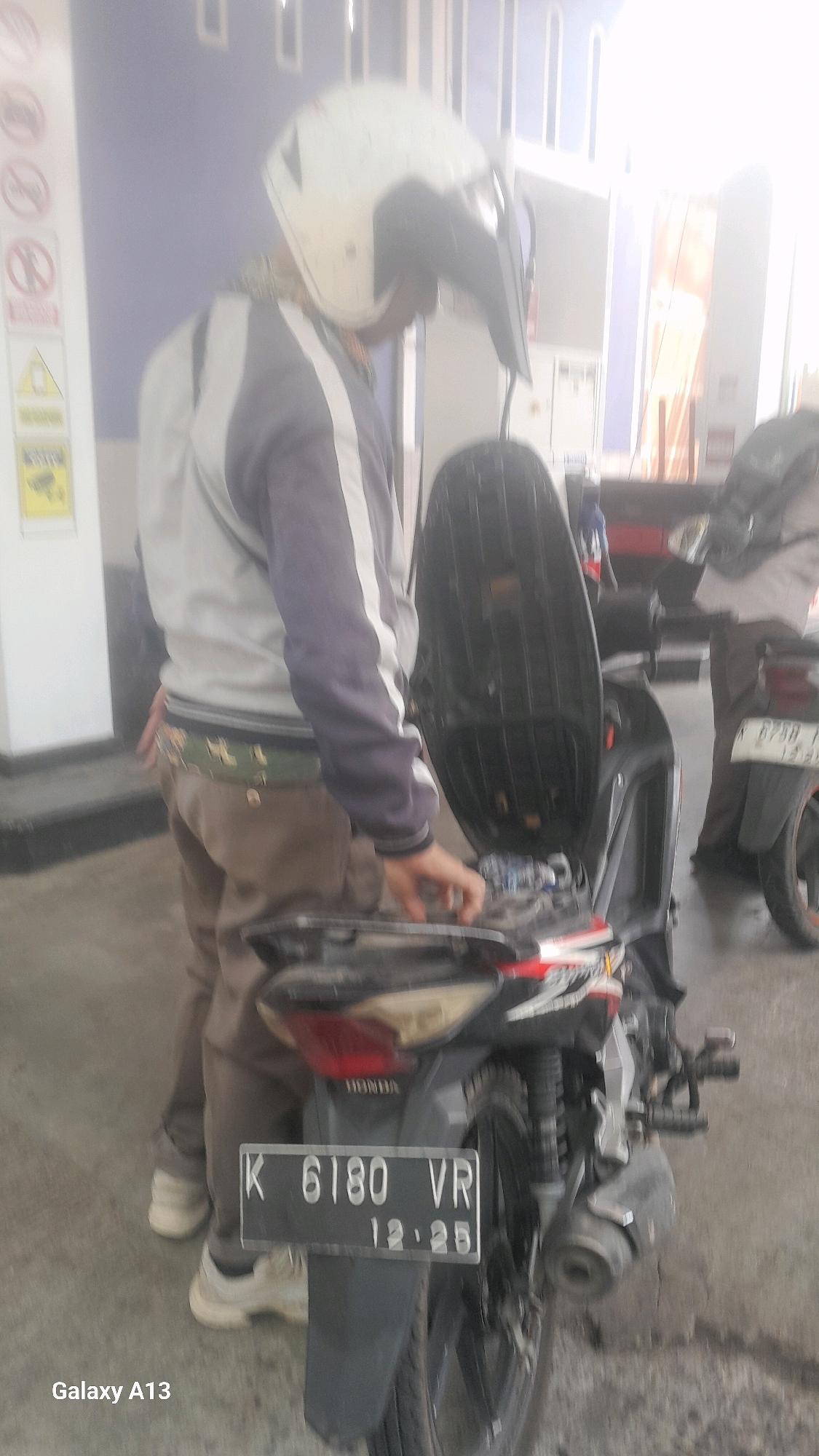 In Tanjunggemuk, Indonesia, a person wearing a white helmet, a grey and dark blue jacket, and brown pants is standing next to a black motorcycle. The motorcycle's seat is open, revealing its interior. A license plate on the rear of the motorcycle reads "K 6180 VR 12.25". The ground is paved, and a wall is visible in the background, along with what appears to be a gas station.