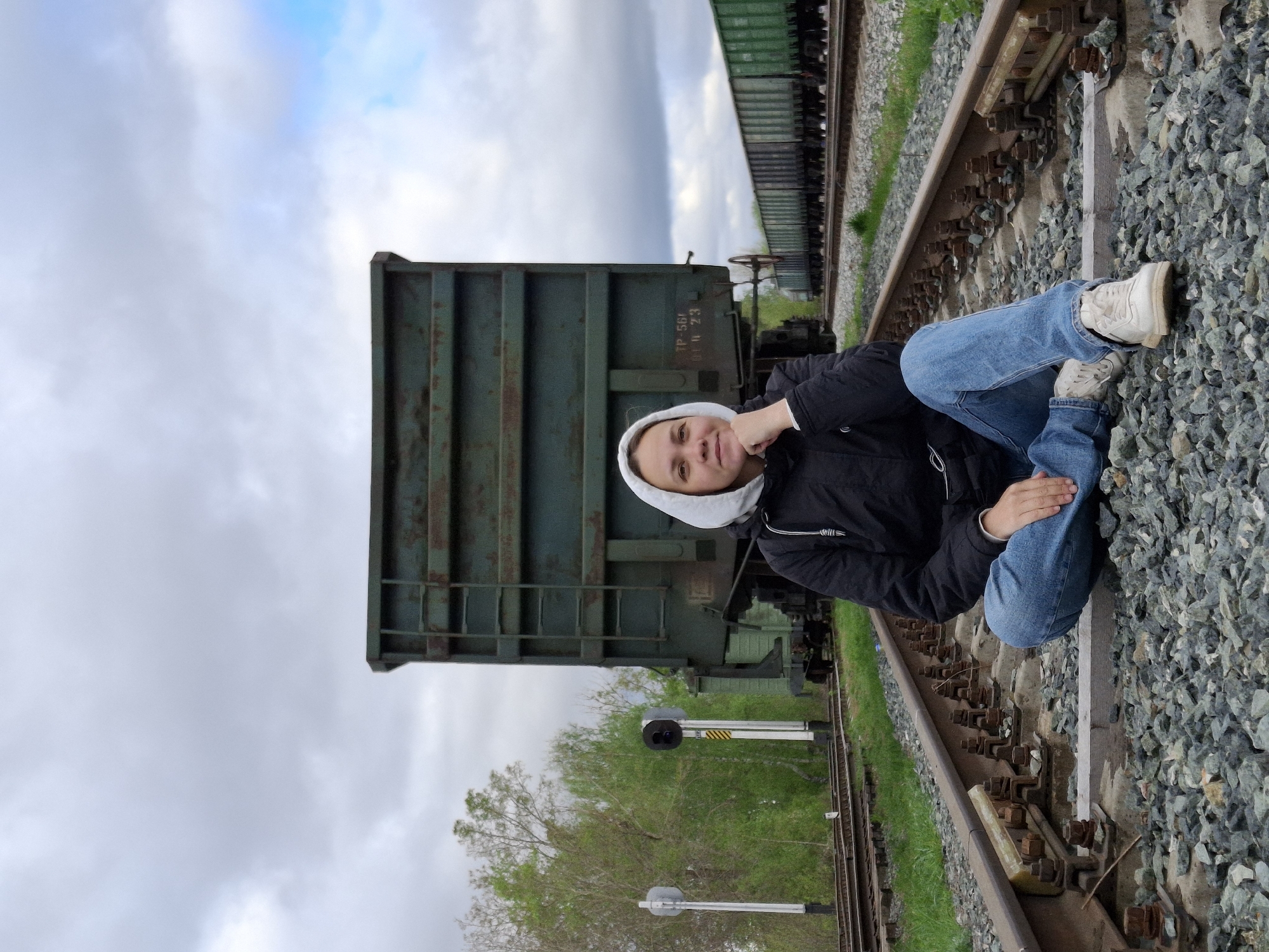 A young woman in a black jacket, blue jeans, and white sneakers is sitting on the gravel next to a railroad track. She is wearing a white hooded sweatshirt. The railroad track has green freight cars on it. In the background, there are trees and railroad signals. The sky is cloudy. The location appears to be near the city of Chaikovsky, Russia based on other visual information.