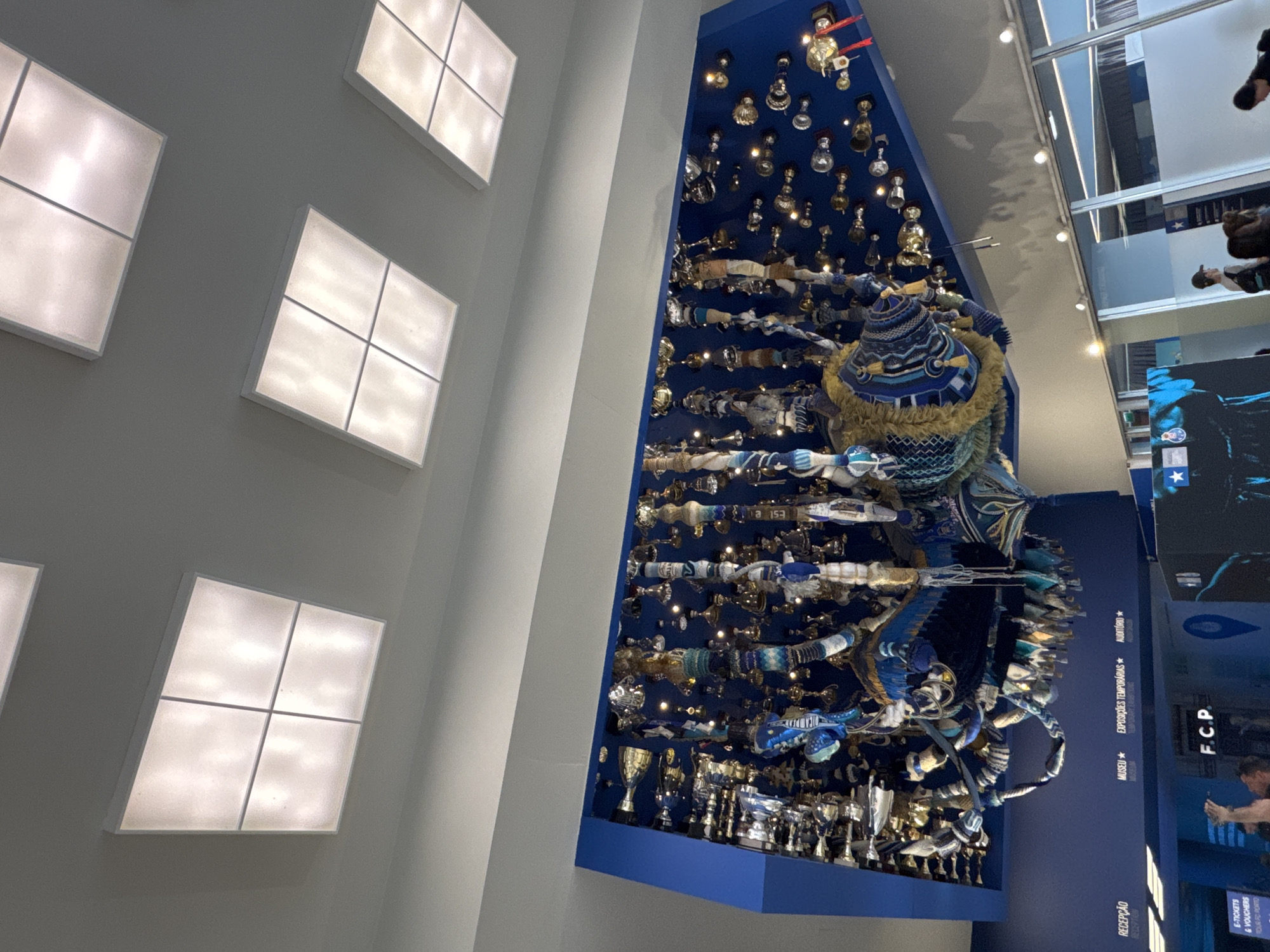 This interior shot showcases an ascending display of artifacts, including trophies, scarves, and other symbolic items, mounted on a tall, blue platform. It seems to reflect the historical journey of a soccer team. The display is topped by a hat-like structure made of blue fabric and golden frills. A series of window-like light fixtures is visible on the wall.
The location is the FC Porto Museum in Porto, Portugal. People are visible as a video.
--- OCR End ---