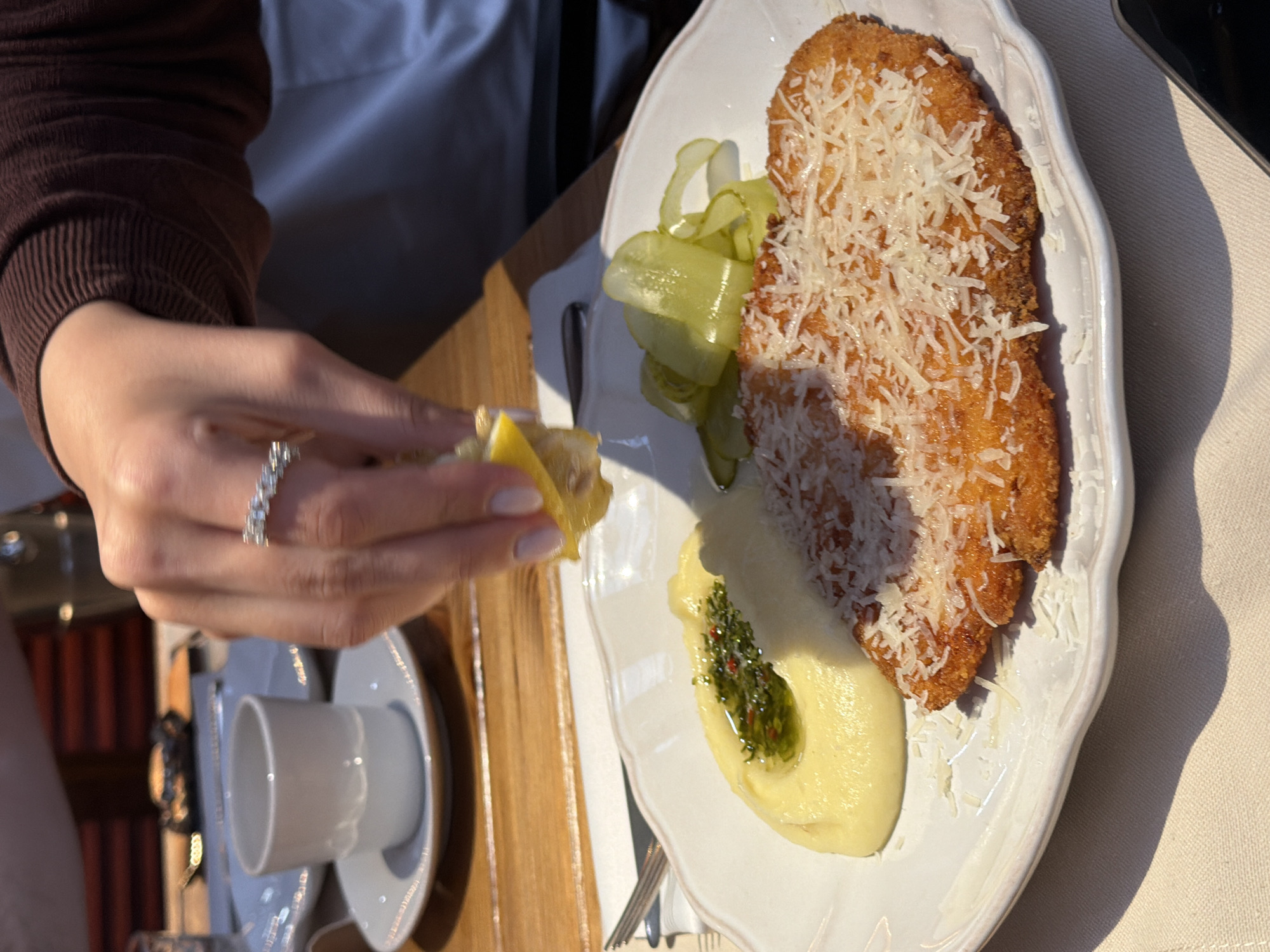 A close-up captures a person squeezing lemon over a plate of food. The dish features a breaded cutlet sprinkled with shredded cheese, accompanied by a dollop of mashed potatoes with a green garnish and slices of pickled cucumber. The person squeezing the lemon wears a brown long-sleeved shirt and an eternity ring on their ring finger. A white teacup and saucer are visible on the wooden table in front of them. The scene is likely set in a restaurant in Yerevan, Armenia, based on the style of food.
