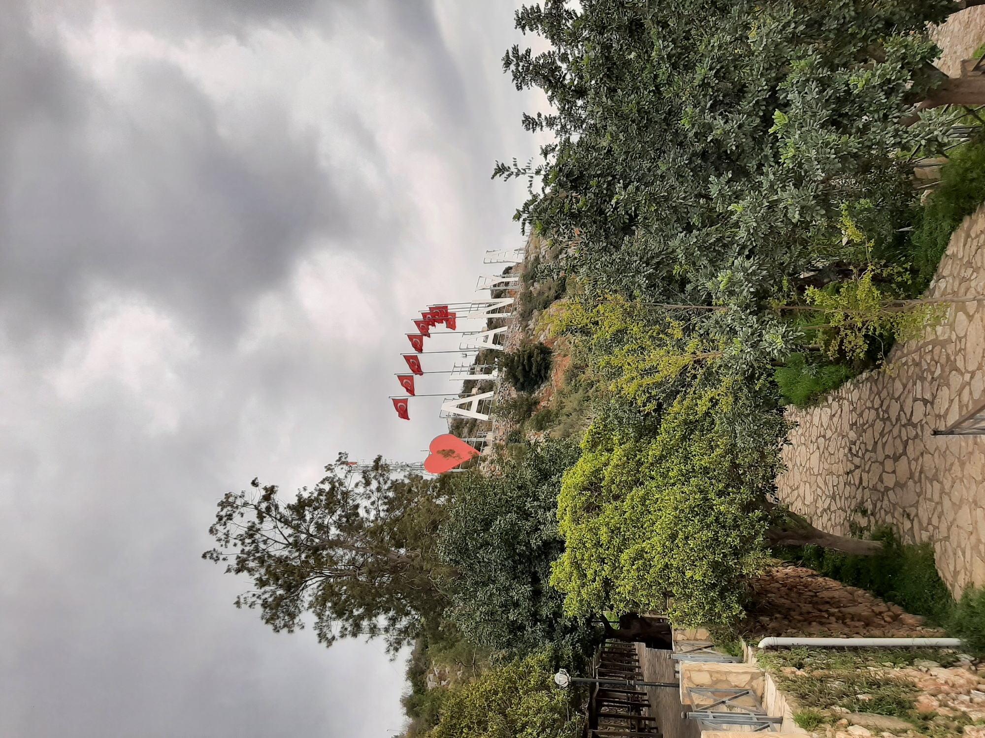 The image depicts a hillside in Alanya, Turkey, with the word "I LOVE ALANYA" spelled out in large white letters. Turkish flags are positioned along the hillside leading up to the sign. A large red heart shape is also visible. Lush green trees and foliage surround the hillside. A stone pathway is on the right. The sky is overcast with grey clouds.