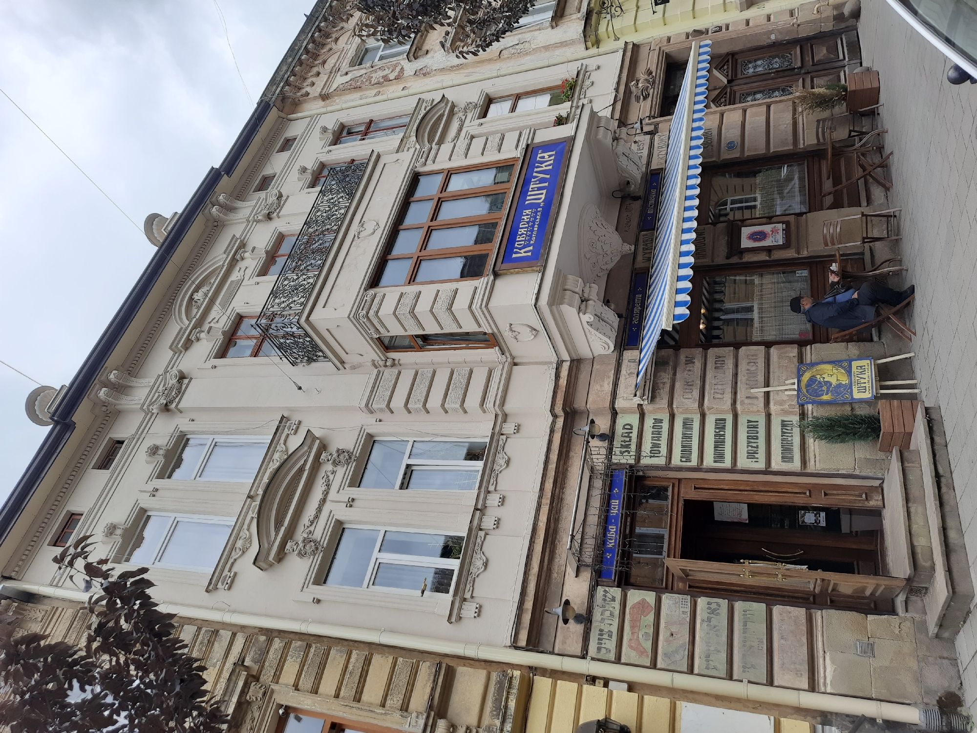 The image depicts a building in Lviv, Ukraine, featuring a facade with classical architectural elements. The building has multiple stories with ornate detailing around the windows and along the roofline. A blue sign on the second story reads "КАВ'ЯРНЯ ШТУКА," indicating a coffee shop. Below this, on the ground floor, is the entrance to the establishment, with an open wooden door and a set of stone steps. A blue and white striped awning covers the storefront windows, under which are tables and chairs set up for outdoor seating. A man wearing a dark jacket and a cap is seated at one of the tables, facing away from the camera. To the right of the entrance, a small evergreen tree is planted in a wooden planter, and a sign with a depiction of a woman and the name "ШТУКА" is placed on an easel. The street is paved with cobblestones. The sky is overcast.