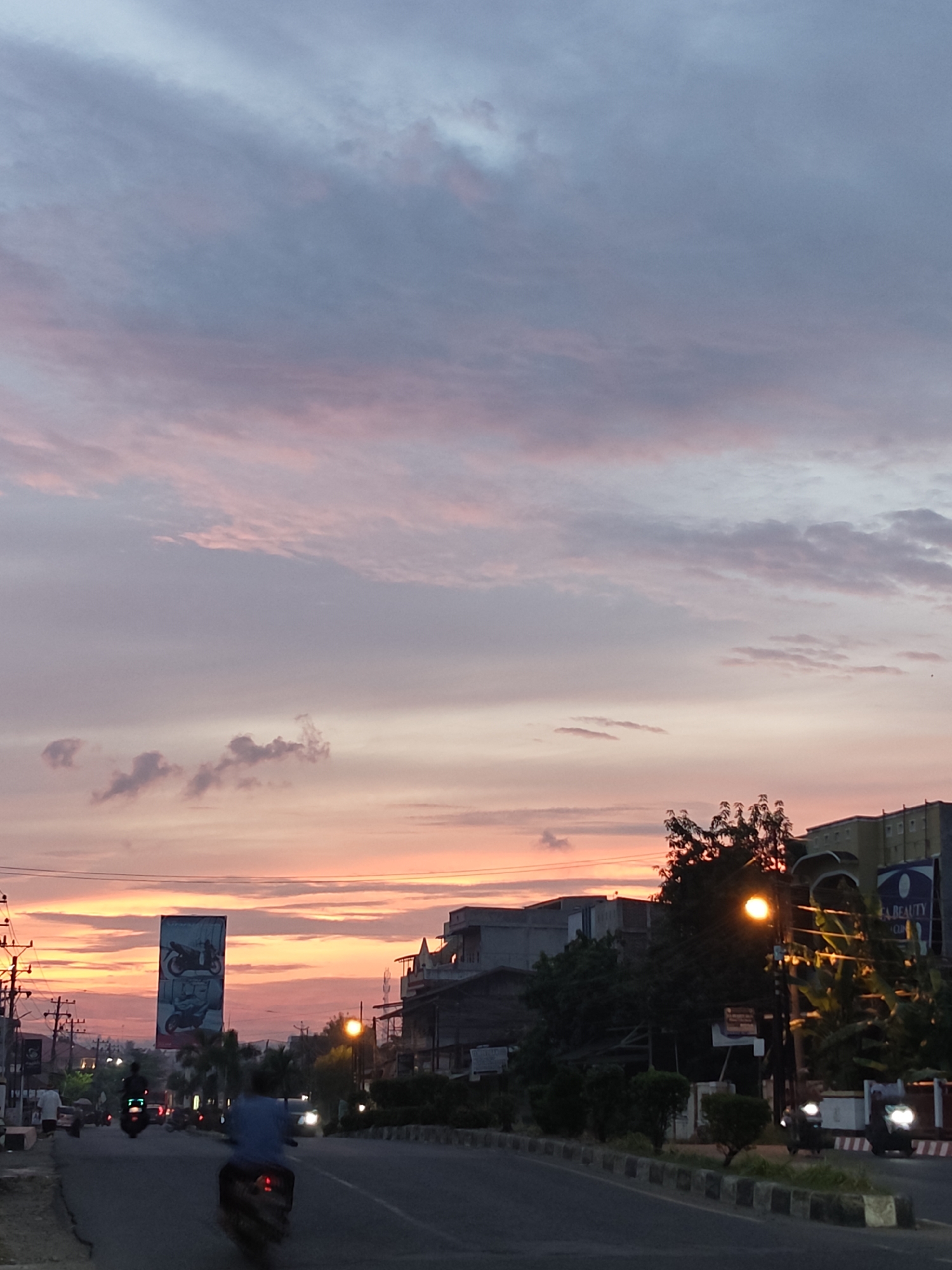 This media file captures a sunset scene in Lhokseumawe, Indonesia. The sky is a gradient of soft pinks, oranges, and blues, with wispy clouds scattered across the expanse. Below the sky, a street is visible, with several motorcycles in motion, suggesting a busy urban environment.
In the foreground, the road curves to the right, bordered by a median strip with concrete barriers and low-lying vegetation. On the left side of the road, utility poles with numerous wires are prominent. Further down the street, a large billboard displays images of two motorcycles. Buildings line the right side of the road, with a mix of architectural styles, including some multi-story structures. A streetlamp emits a warm glow, illuminating the immediate surroundings.
The overall mood of the image is serene, with the vibrant colors of the sunset creating a peaceful atmosphere. The presence of vehicles and buildings indicates an inhabited area, likely a city or town. The time of day is clearly sunset, given the sky's colors and the artificial lighting beginning to illuminate. While no specific event is depicted, the scene conveys a sense of daily life transitioning into evening. There is a visible sign that reads "BEAUTY" on one of the buildings on the right.
