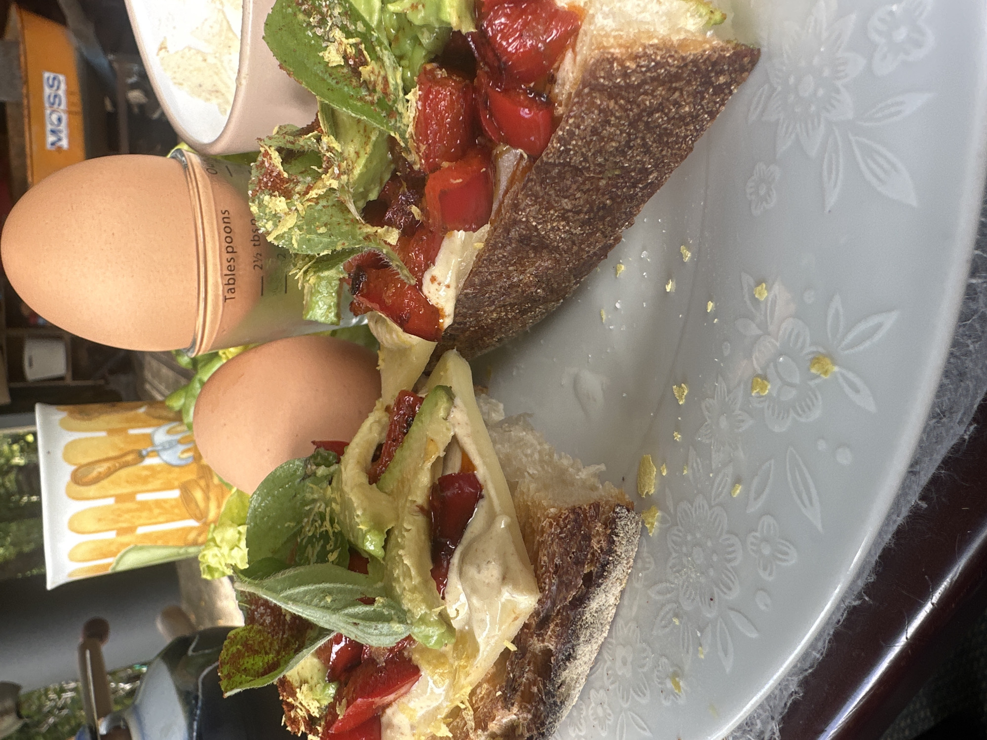 The image shows a close-up of an open-faced sandwich on a floral embossed white plate. The sandwich is made with a slice of dark, crusty bread topped with a creamy spread, sliced avocado, roasted red peppers, and fresh green herbs. There are also two boiled eggs in the background, one in a small glass measuring cup, and a ceramic mug with a yellow and brown pattern. A small orange wheelbarrow labeled "MOSS" is visible in the far background. The scene appears to be outdoors, possibly on a patio or balcony, given the natural light and the presence of greenery. The overall setting suggests a casual meal, likely breakfast or lunch. There is no indication of the city of Thora, Australia in the image.