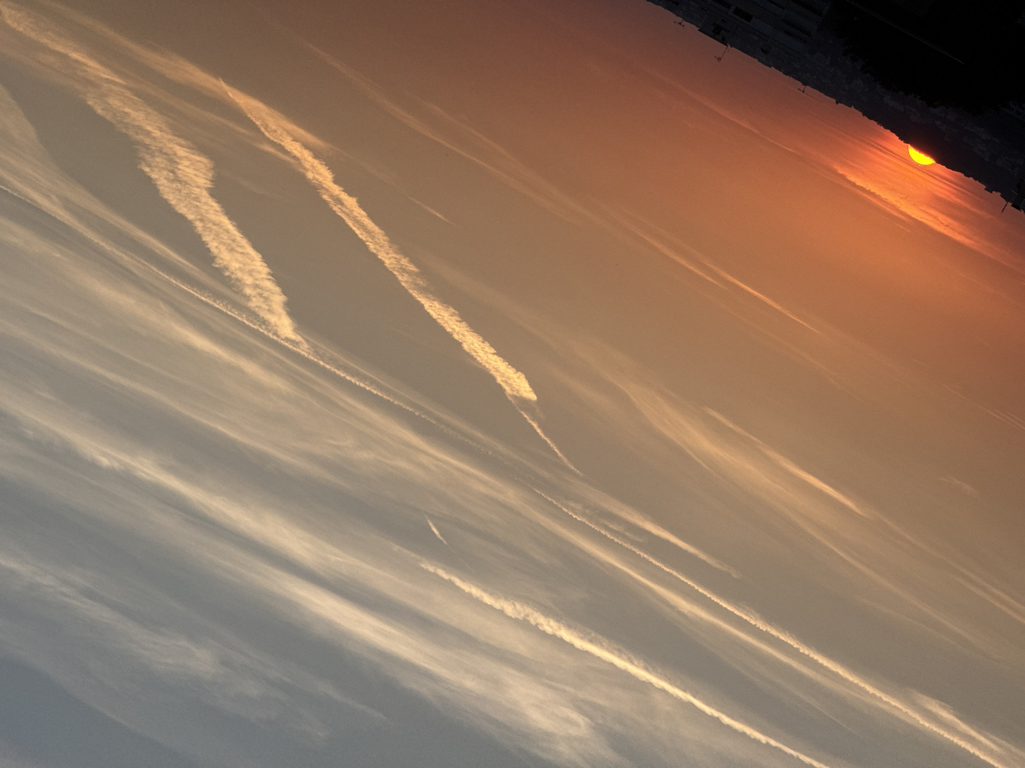 This is a high-angle, wide shot of a sunset over a city. The sun is a bright orange semi-circle peeking over a silhouetted hillside covered with buildings. The sky is filled with diagonal lines of wispy clouds, some appearing as contrails from airplanes, catching the golden and orange light of the setting sun. The bottom of the image is dominated by the darker silhouettes of buildings and trees in the foreground, with the sun illuminating the sky above them. The perspective is tilted, giving the impression of a diagonal landscape. The location is listed as Unknown, Unknown.