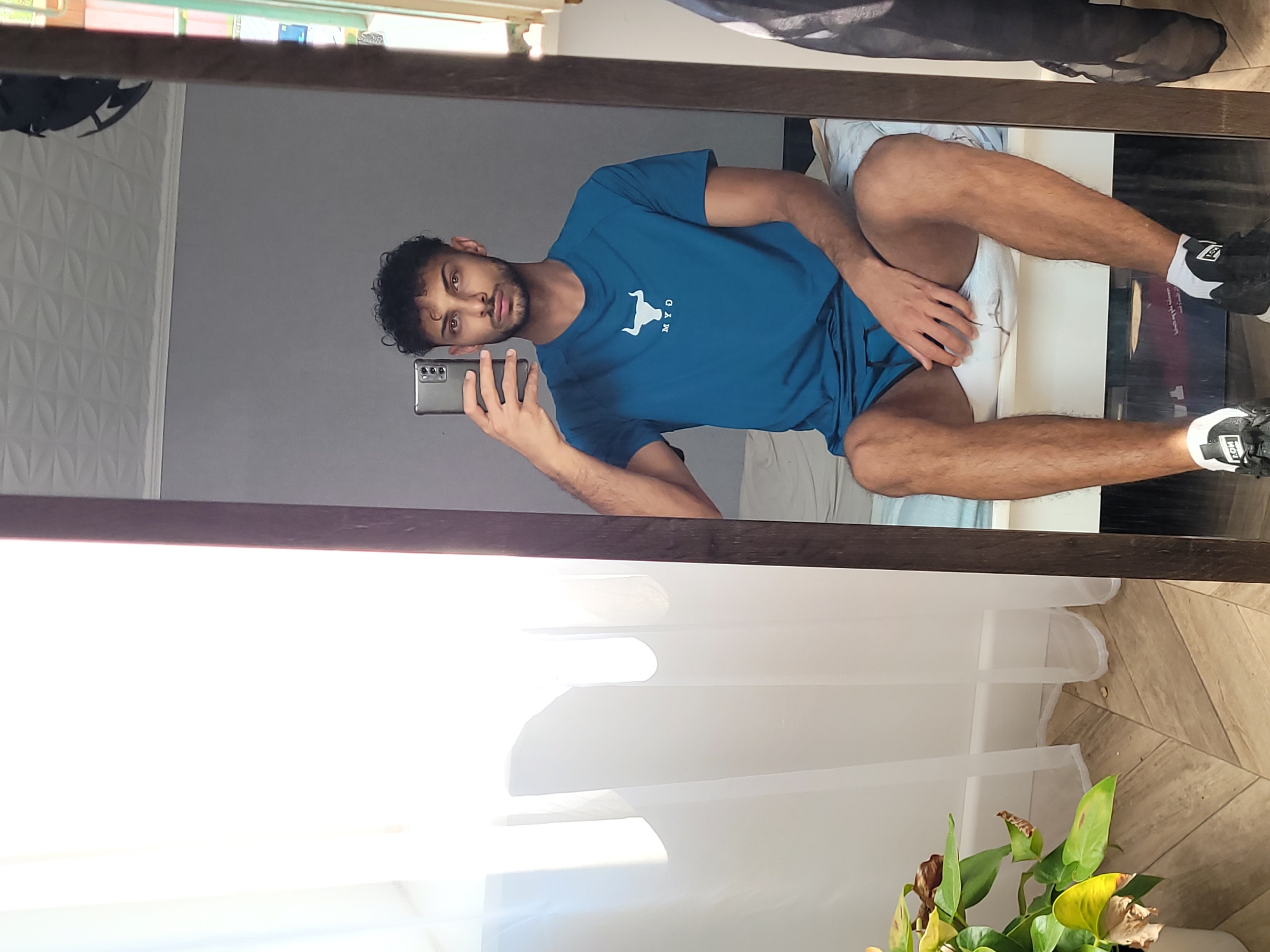 In a room with light-colored walls and a textured ceiling, a young man with curly dark hair and a beard is taking a selfie in a full-length mirror. He is wearing a teal t-shirt with a white bull logo and the letters "MYO" below it, and matching teal shorts. He is sitting on a bed covered with a white duvet. The mirror frame is dark brown. To the left of the mirror, sheer white curtains hang over a window, letting in bright sunlight. A potted plant with green and yellow leaves is visible in the lower left corner of the frame. The floor has a herringbone pattern of light brown tiles. To the right of the man, a black garment, possibly clothing, is draped over something not fully visible. The overall impression is of a casual indoor setting, potentially a bedroom. The location of Givors, France is not discernible from the image.