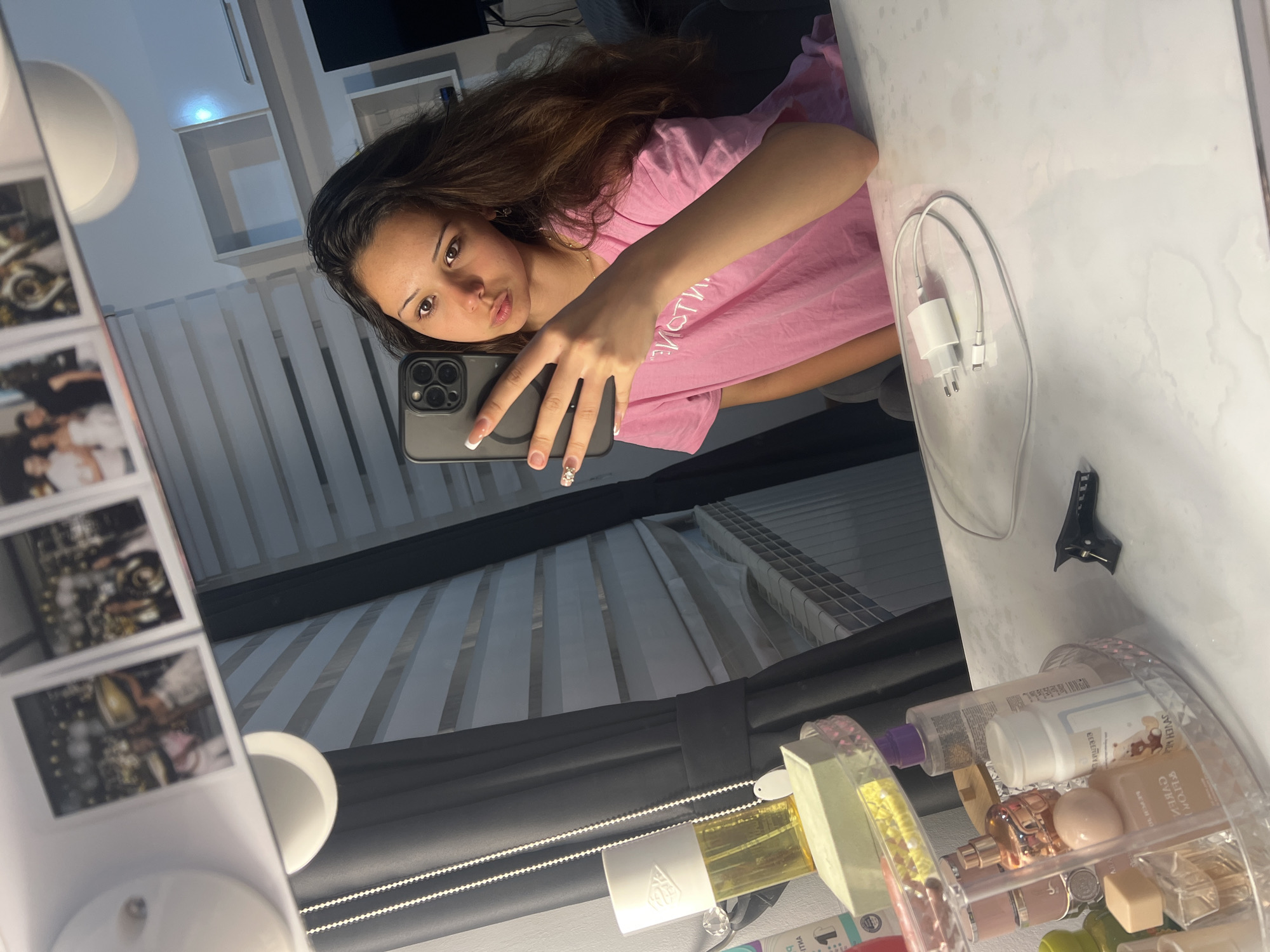 A young woman is depicted in a mirror selfie, capturing her reflection in what appears to be a vanity or dressing area. She has long, dark brown hair and is wearing a pink t-shirt with partially visible text that reads "MOTIV". Her left hand, adorned with French-tip manicured nails, holds an iPhone with a triple camera system, making a peace sign gesture. She gazes directly into the camera with a slight pout on her lips.
The reflection reveals a bright, indoor setting. To the left of the woman, a white, possibly round lamp or light fixture is visible, alongside a stand displaying multiple small, square framed photos, which are blurred but appear to show various scenes or individuals. Behind her, a window with horizontal blinds provides a backdrop, while white shelves are mounted on a light-colored wall.
On the reflective surface to her right, a white charging brick with a two-prong European-style plug is connected to two white cables. Below it, a clear acrylic organizer holds various cosmetic and fragrance items, including bottles with visible text such as "ELEMENTS," "3CE" (a Korean beauty brand), and partially "DIOR." A small black clip is also seen on the surface. The mirror itself shows some water stains or marks. The scene suggests a personal space, likely a bedroom or dressing room, with a soft, artificially lit atmosphere.