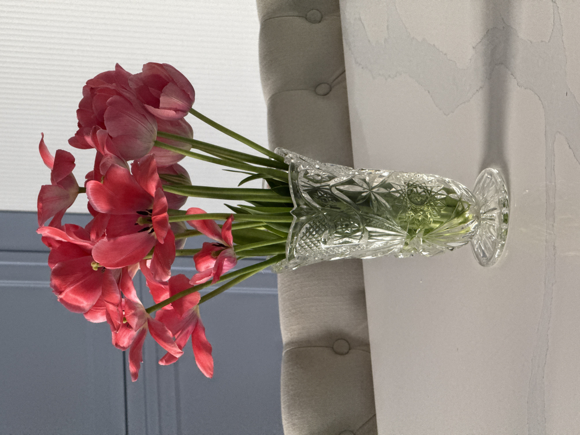A clear, decorative vase filled with water and a bouquet of pink tulips sits on a white surface. A grey upholstered surface with button detailing is partially visible behind the vase. The setting appears to be indoors. The image does not contain any information to suggest a location in Moscow, Russia, or any city.