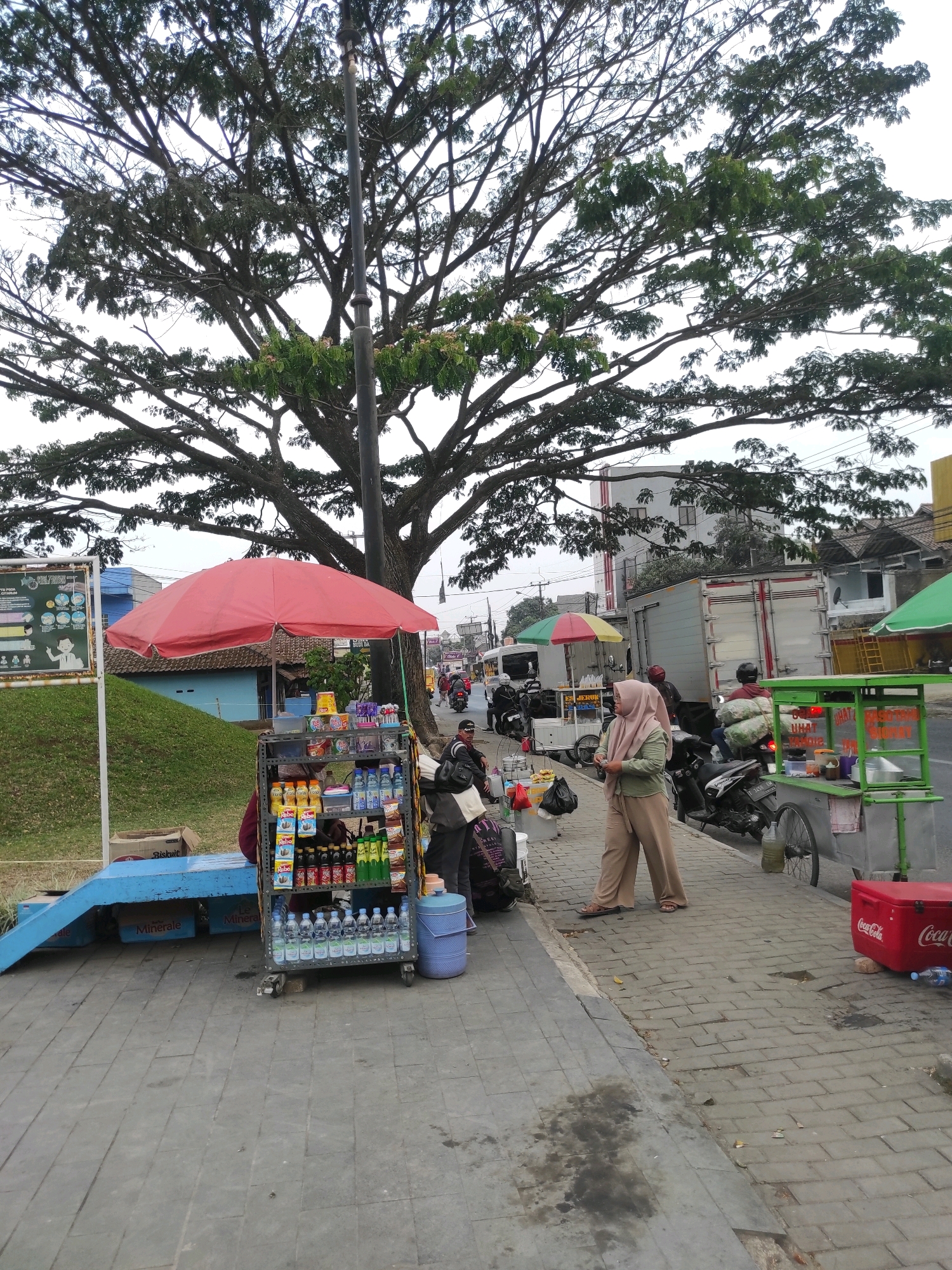 The image depicts a street scene in Ciburuy, Indonesia, dominated by a large tree with extensive branches. In the foreground, a sidewalk is lined with various stalls and parked vehicles. A woman wearing a light brown hijab and loose-fitting beige pants stands near a food cart. Beside her, a small red Coca-Cola branded cooler is placed on the ground. A street vendor's cart is visible, stocked with bottled drinks and other items, covered by a red umbrella. Further down the sidewalk, another cart with a green canopy is set up. Several motorcycles are parked, and a large truck is visible in the background, along with buildings and other commercial structures. The ground is paved with interlocking bricks.